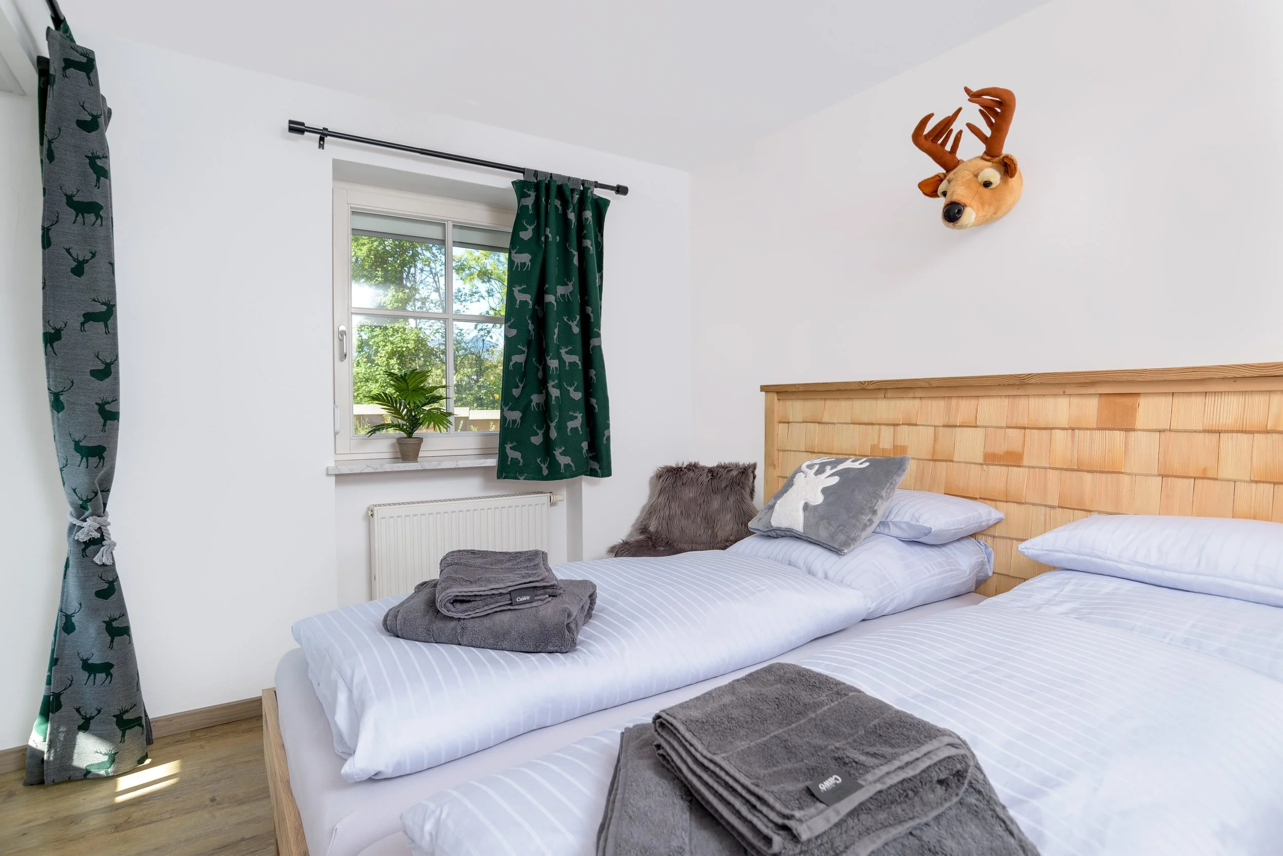 Berg Fux Ferienhaus Allgaeu Panorama Schlafzimmer mit Betten, grauen Handtüchern, Kissen, Fenster mit dschungelartigen Vorhängen, einem grünen Pflanze, einer Hirschkopf-Wanddekoration.