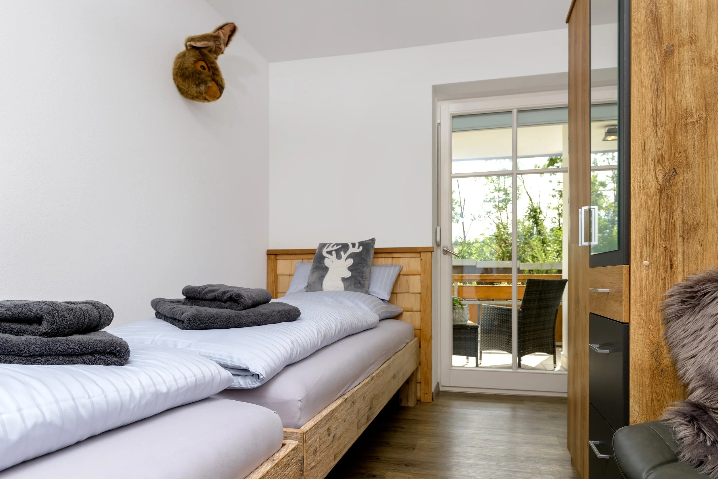 Berg Fux Ferienhaus Allgaeu Panorama Ein Schlafzimmer mit einem Holzbett, weißen Bettwäsche, mehreren grau Handtüchern und einem Kissen mit Hirschmotiv. Es gibt eine große Fensterfront mit Blick nach draußen, einen Kleiderschrank