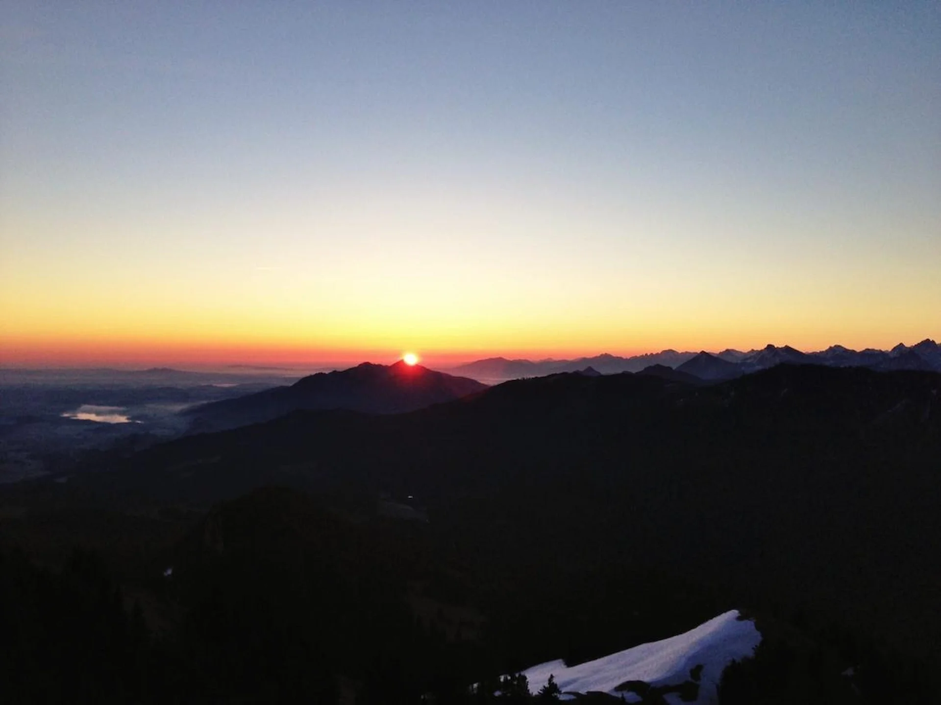 Berglandschaft bei Sonnenaufgang mit hell erleuchtetem Himmel und Bergen im Hintergrund, einige Schneeflächen im Vordergrund. Berg Fux Ferienhaus + Wohnungen