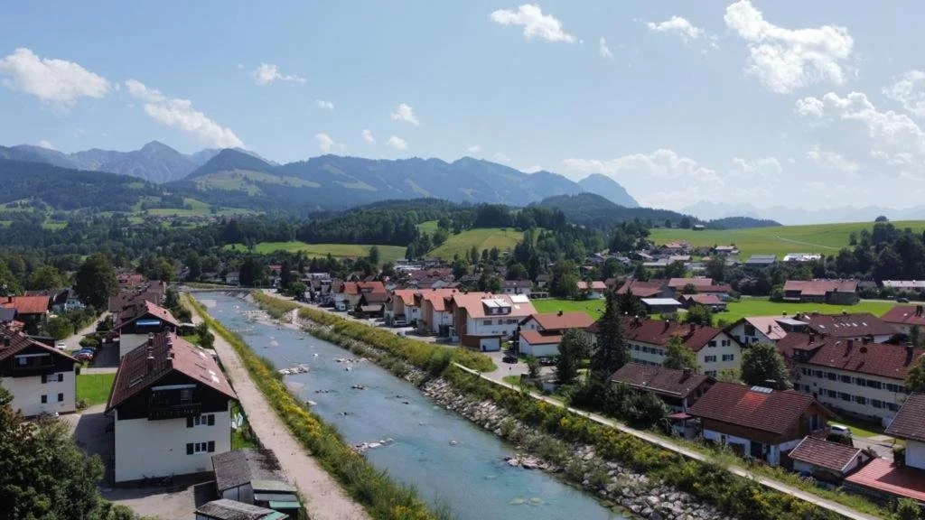 Berg Fux Sonthofen mit Fluss, Häuser mit roten Dächern, grüne Hügel und Berge im Hintergrund, blauer Himmel mit Wolken. Ausblick vom Ferienhaus Berg Fux Fluss Ostrach