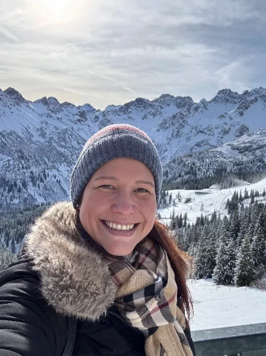Eine lachende Frau mit Mütze, Schal und Winterjacke vor verschneiten Bergen und Tannen im Winter. Berg Fux Ferienhaus + Wohnungen Stefanie Hilgers Inhaberin