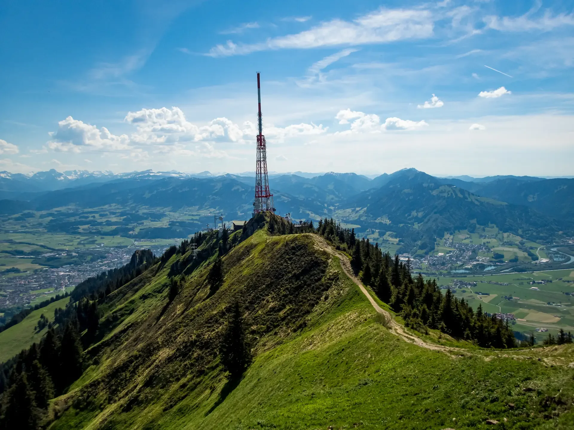 Grüne Berglandschaft mit Wanderpfad im Vordergrund, Funkmast auf Gipfel, Täler und Berge im Hintergrund, blauer Himmel mit Wolken und Flugzeugspur. Berg Fux Ferienhaus + Wohnungen