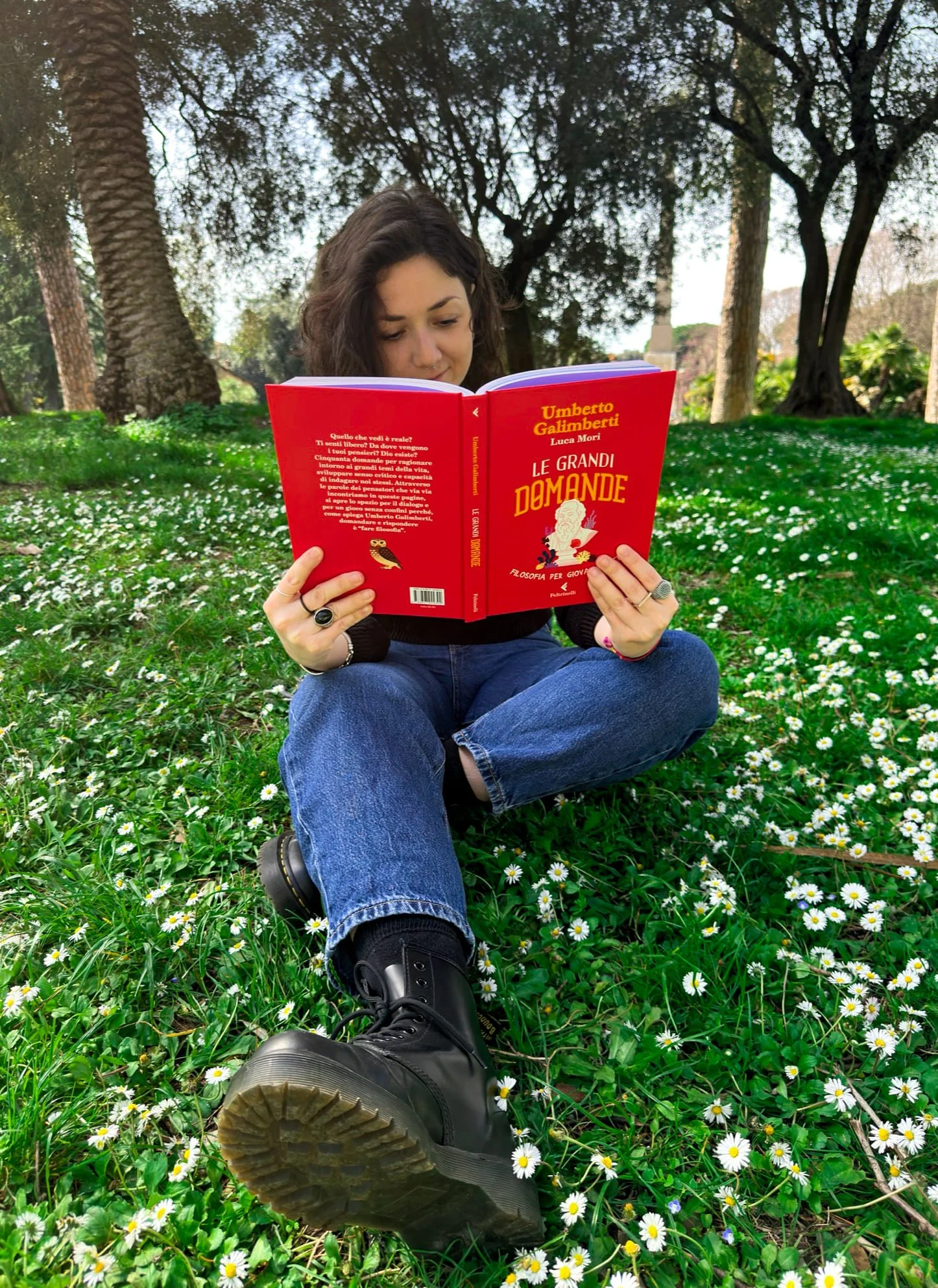 Cristina Damiani. Capelli ricci, abbigliamento casual, seduta su un prato ricoperto di margherite, mentre legge il libro illustrato "Le grandi domande" di Umberto Galimberti ed edito da Feltrinelli. Sullo sfondo ci sono alberi e un cielo sereno.