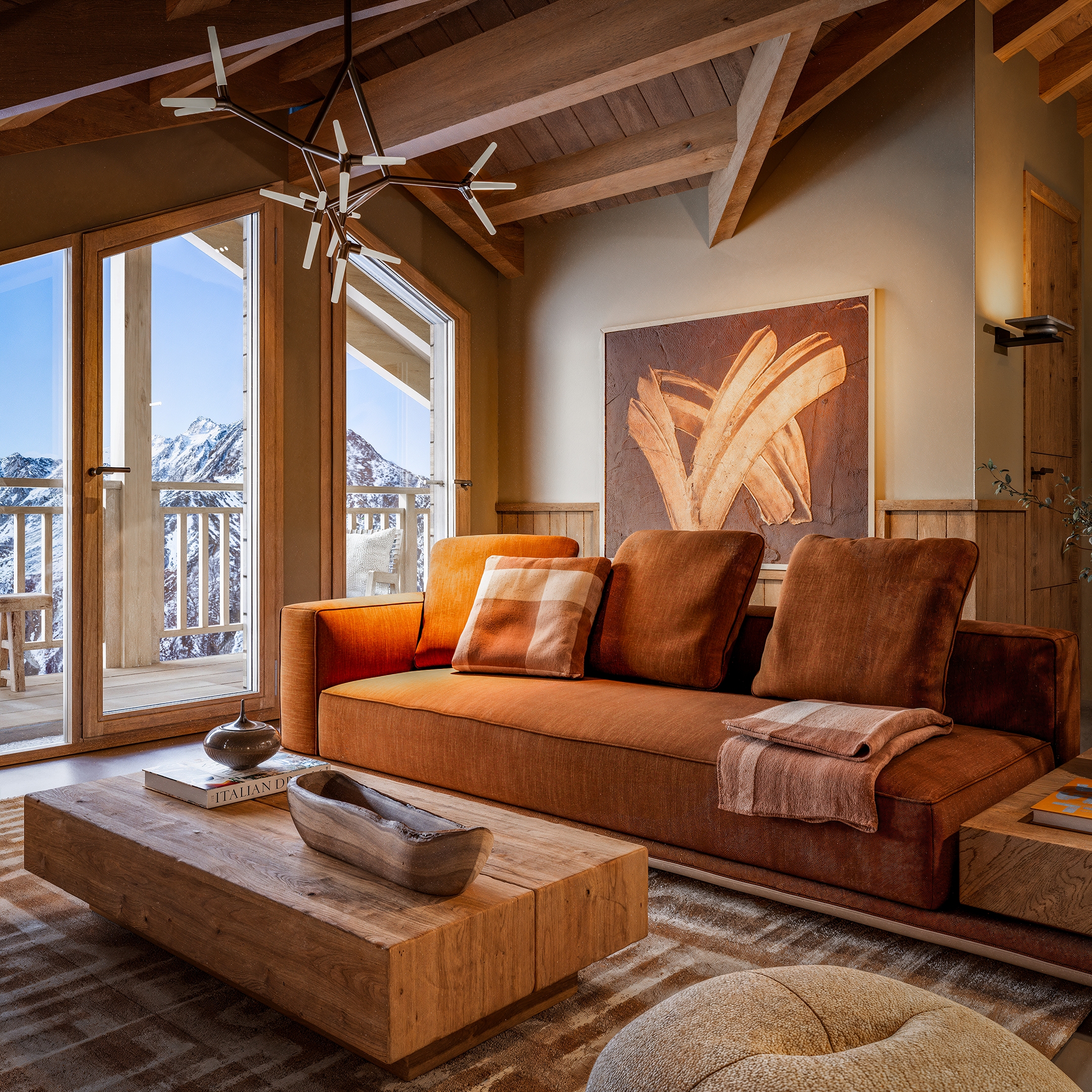 Interieur van een houten huis met grote ramen, uitzicht op besneeuwde bergen, een moderne hanglamp, een kunstwerk en een houten salontafel met boeken, een bank met oranje en geruite kussens, en een vloerkleed.