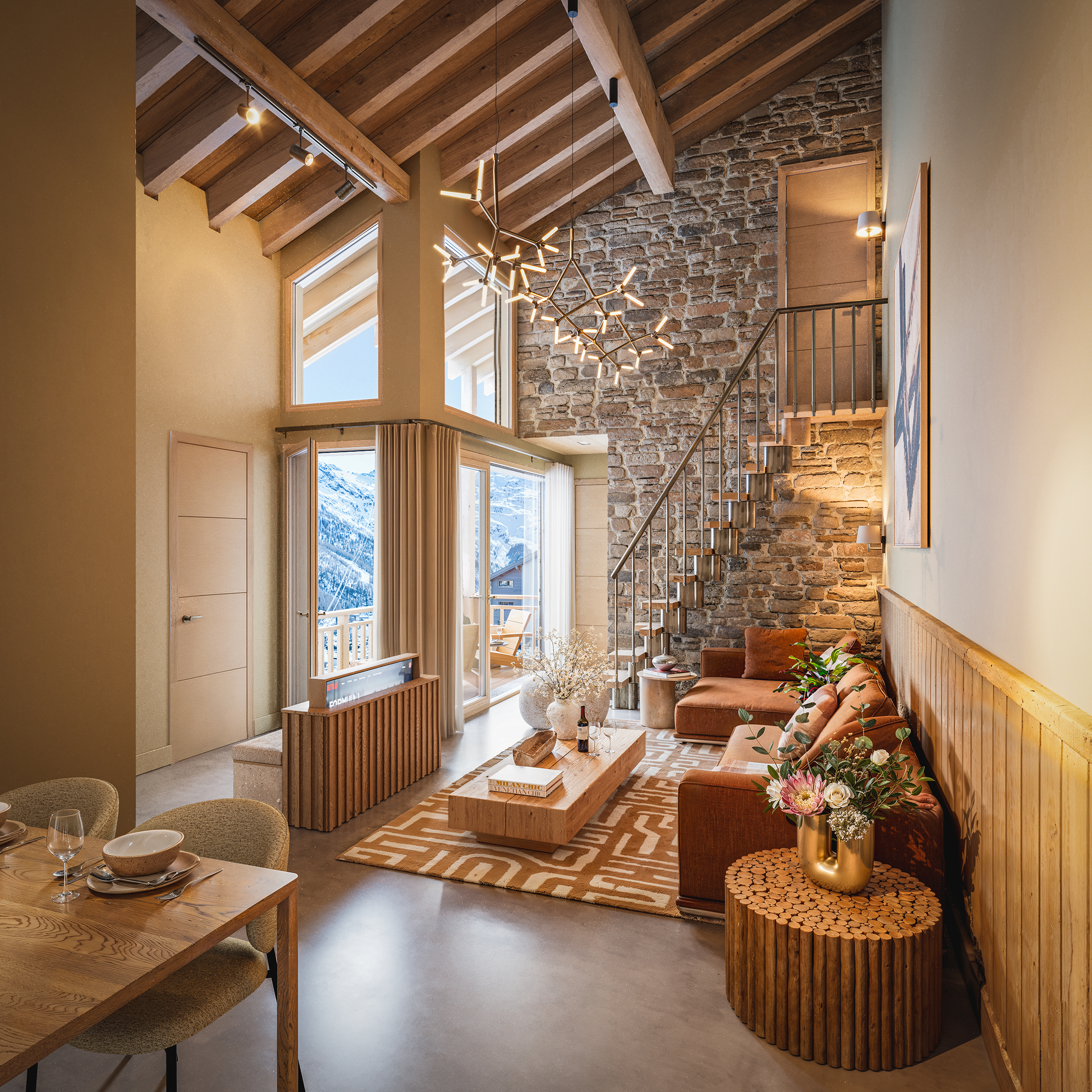 Interieur van een woonkamer met een houten plafond, een stenen muur, grote ramen met uitzicht op een berglandschap, een moderne kroonluchter, een bank met kussens en een salontafel met bloemen en een glas wijn.