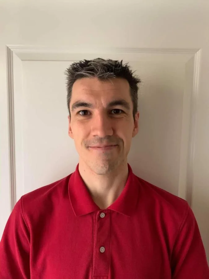 Man with short dark hair and light skin wearing a red polo shirt, standing in front of a white door.