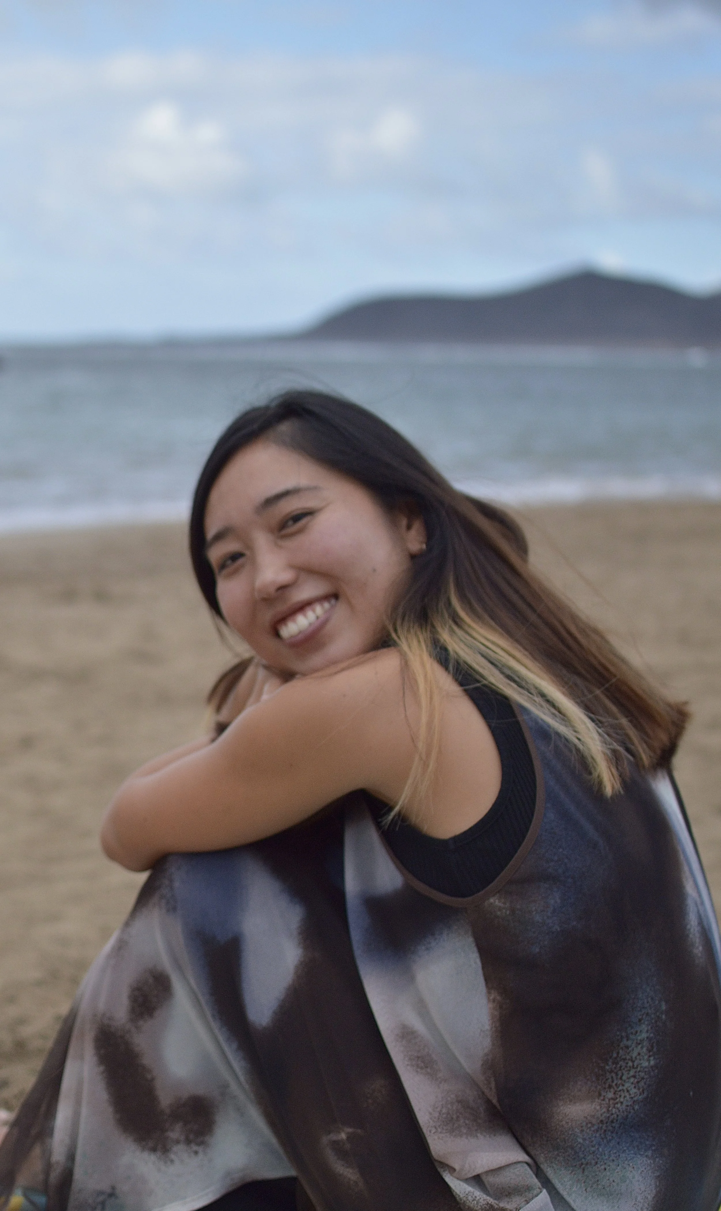 Mujer asiática sentada en la playa mirando hacia la cámara y sonriendo