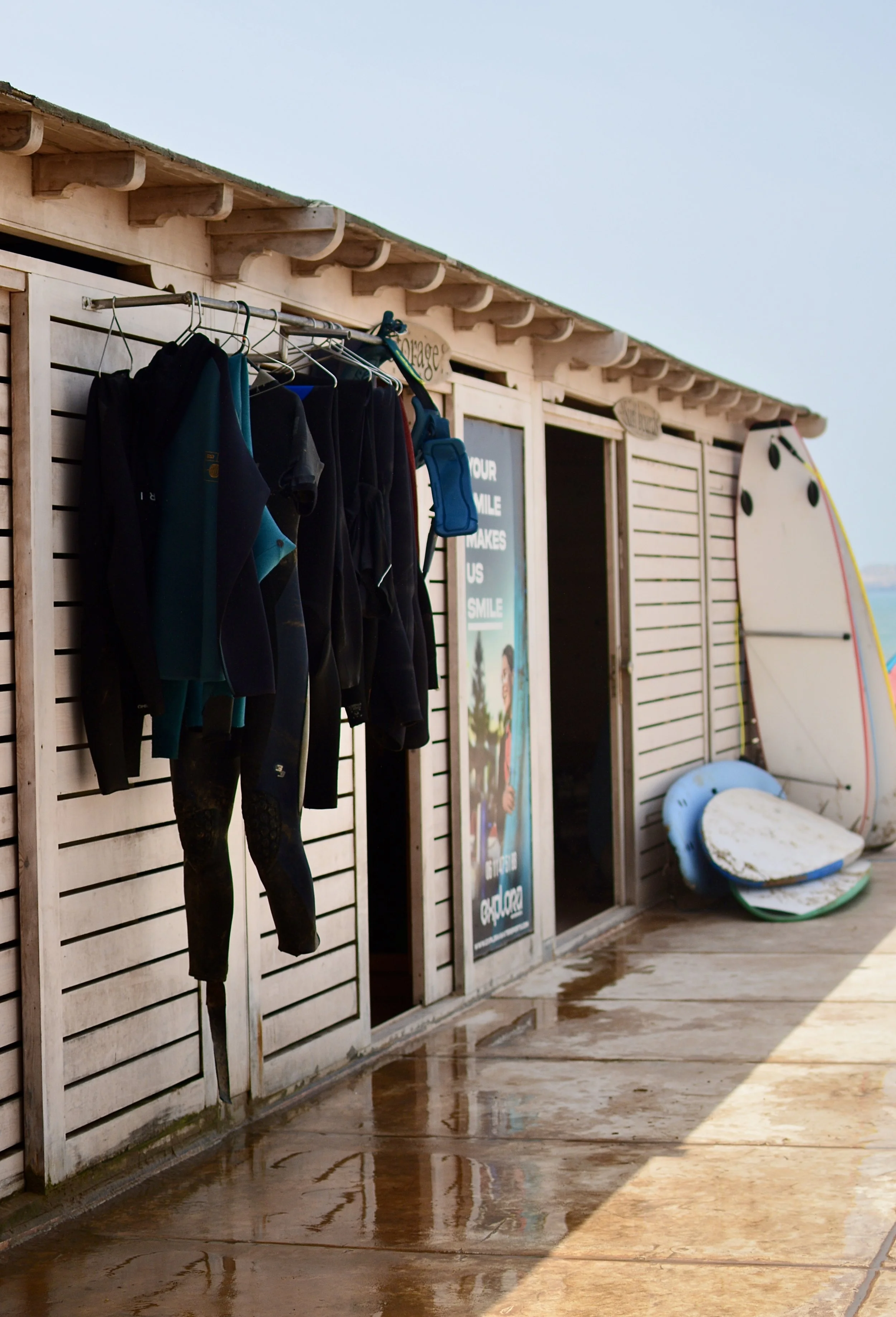 Caseta de una escuela de deportes acuáticos con varias tablas de surf apoyadas y neoprenos colgados en perchas