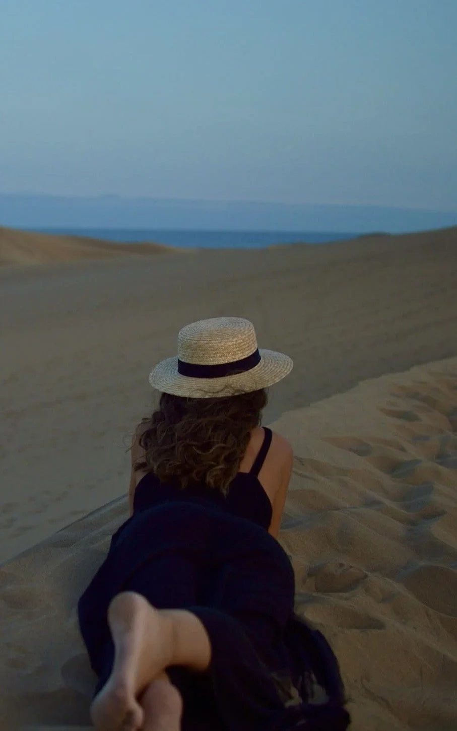 Mujer en unas dunas de la playa tumbada con un sombrero y un vestido negro observando el horizonte