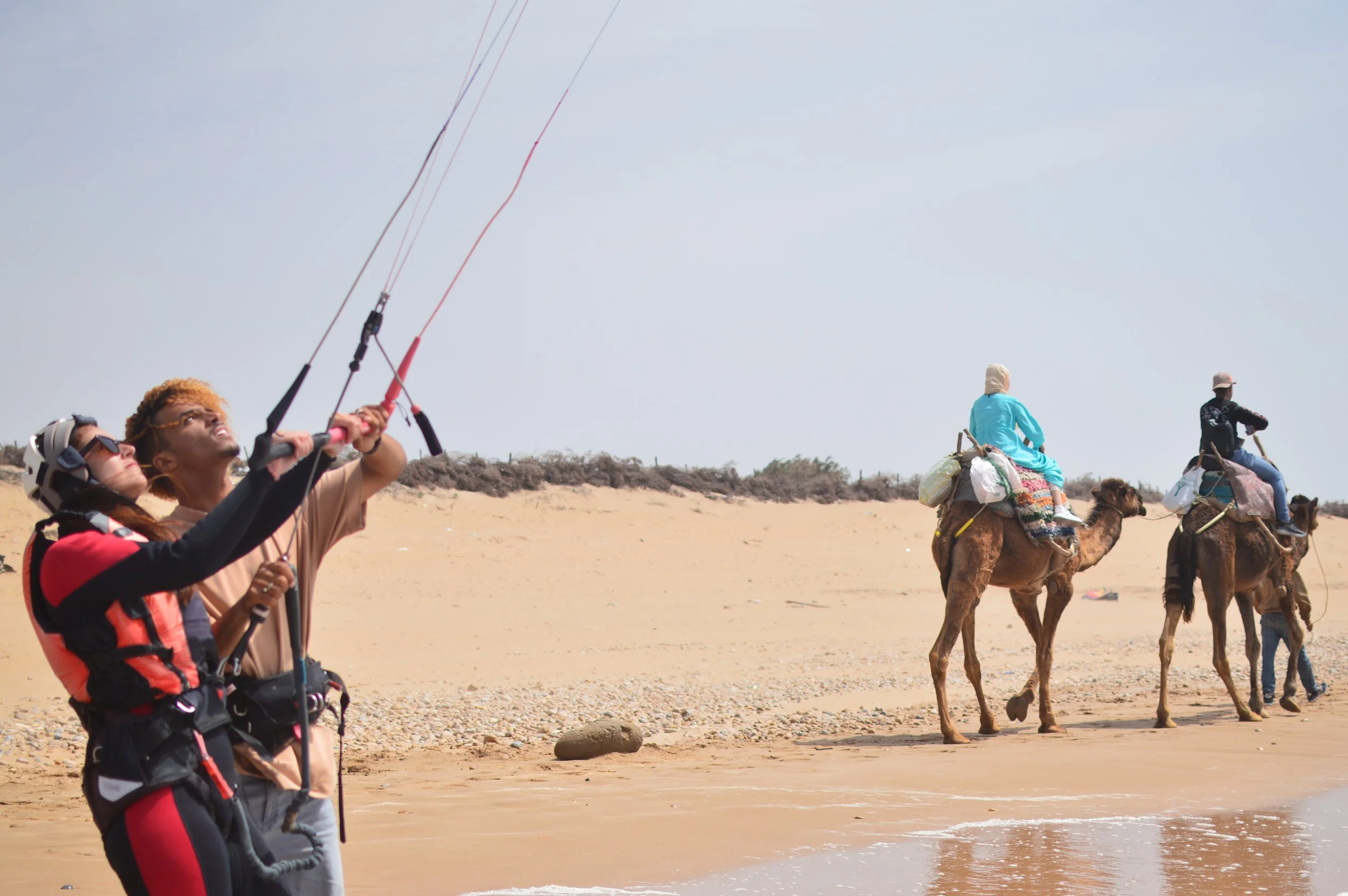 Una mujer aprendiendo a practicar kitesurf junto con su profesor en la playa, con camellos paseando de fondo