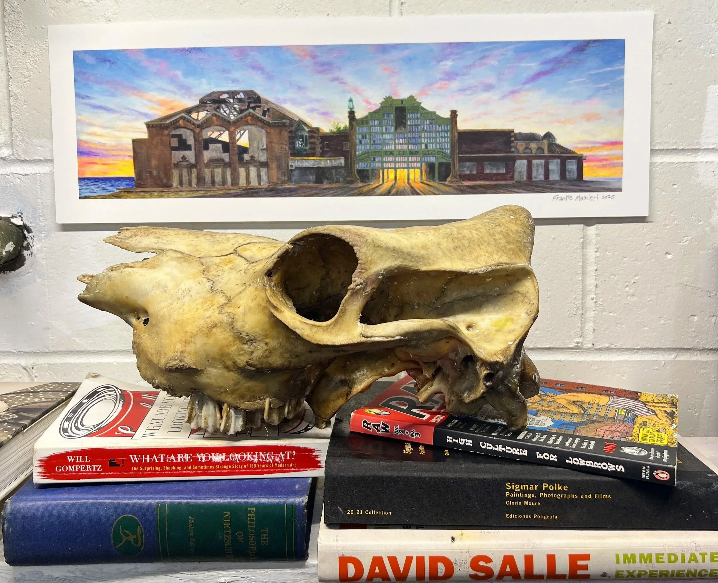 A zebra skull placed on top of a stack of books with a colorful modern artwork of an industrial building and sunset in the background on the wall.