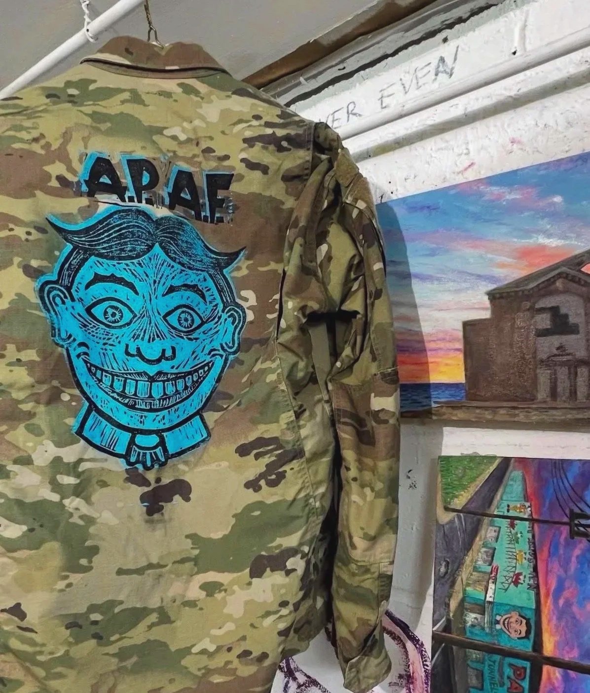 A camouflage military jacket hangs on a wall, with a colorful embroidered patch of a smiling face and the text 'ARAF' on the back. Behind it, there are paintings of a sunset and a cityscape.