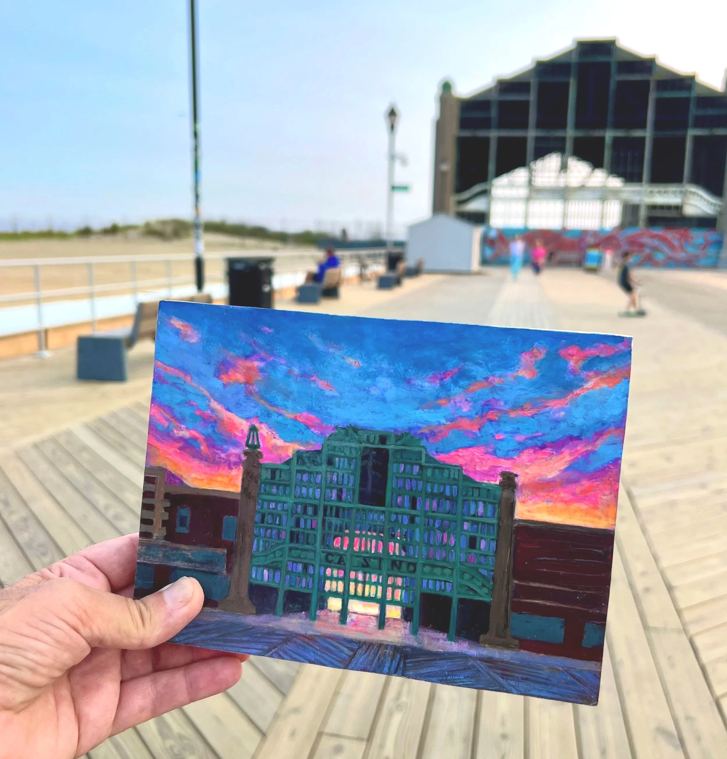Hand holding a colorful painting of a building and sunset sky at a boardwalk.