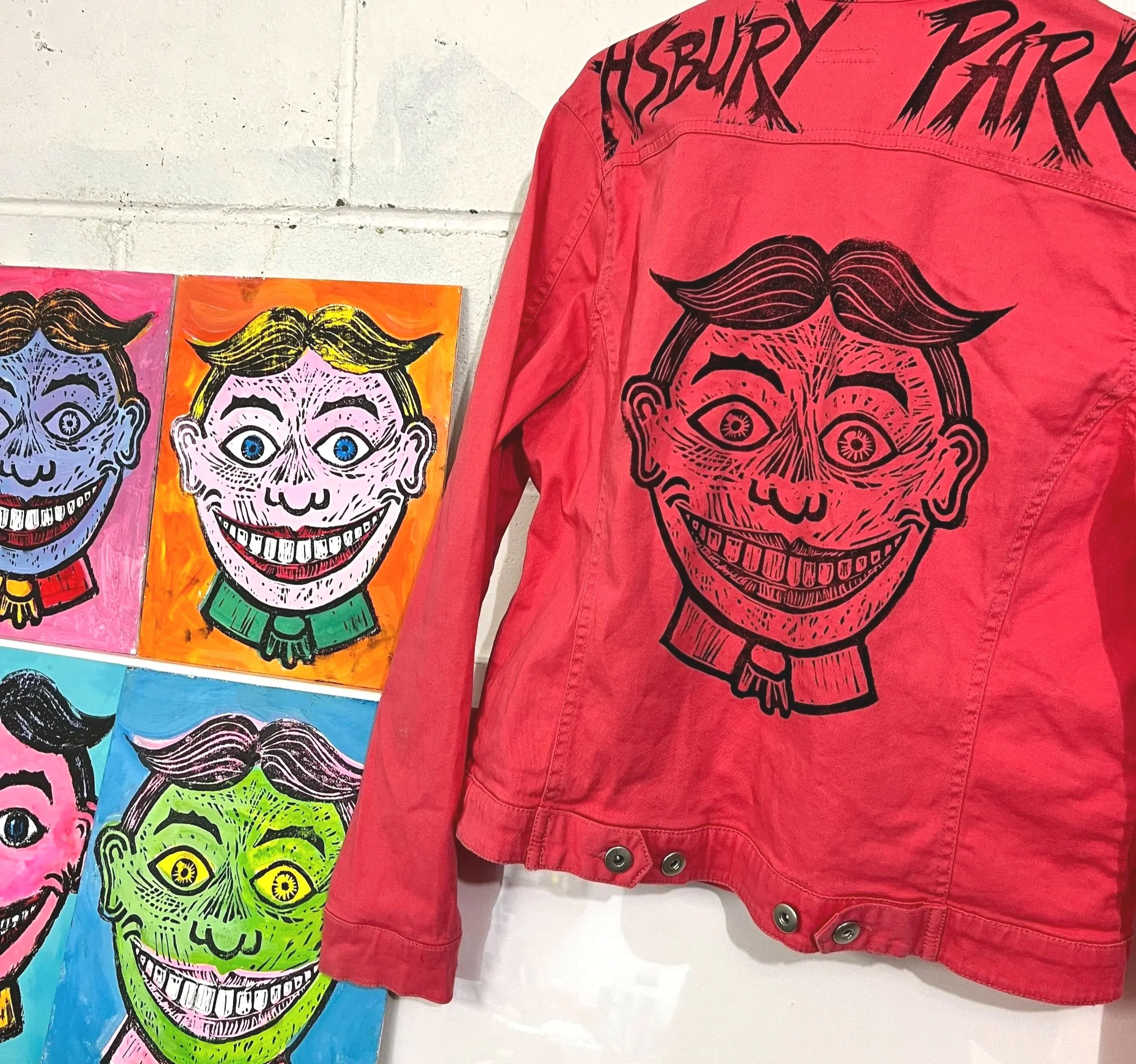 A red jacket hanging on a wall with a large cartoon-like face of a smiling man with styled hair, drawn in black lines. To the left, four colorful paintings of the same face in different colors—pink, orange, blue, and green—are displayed on the wall.