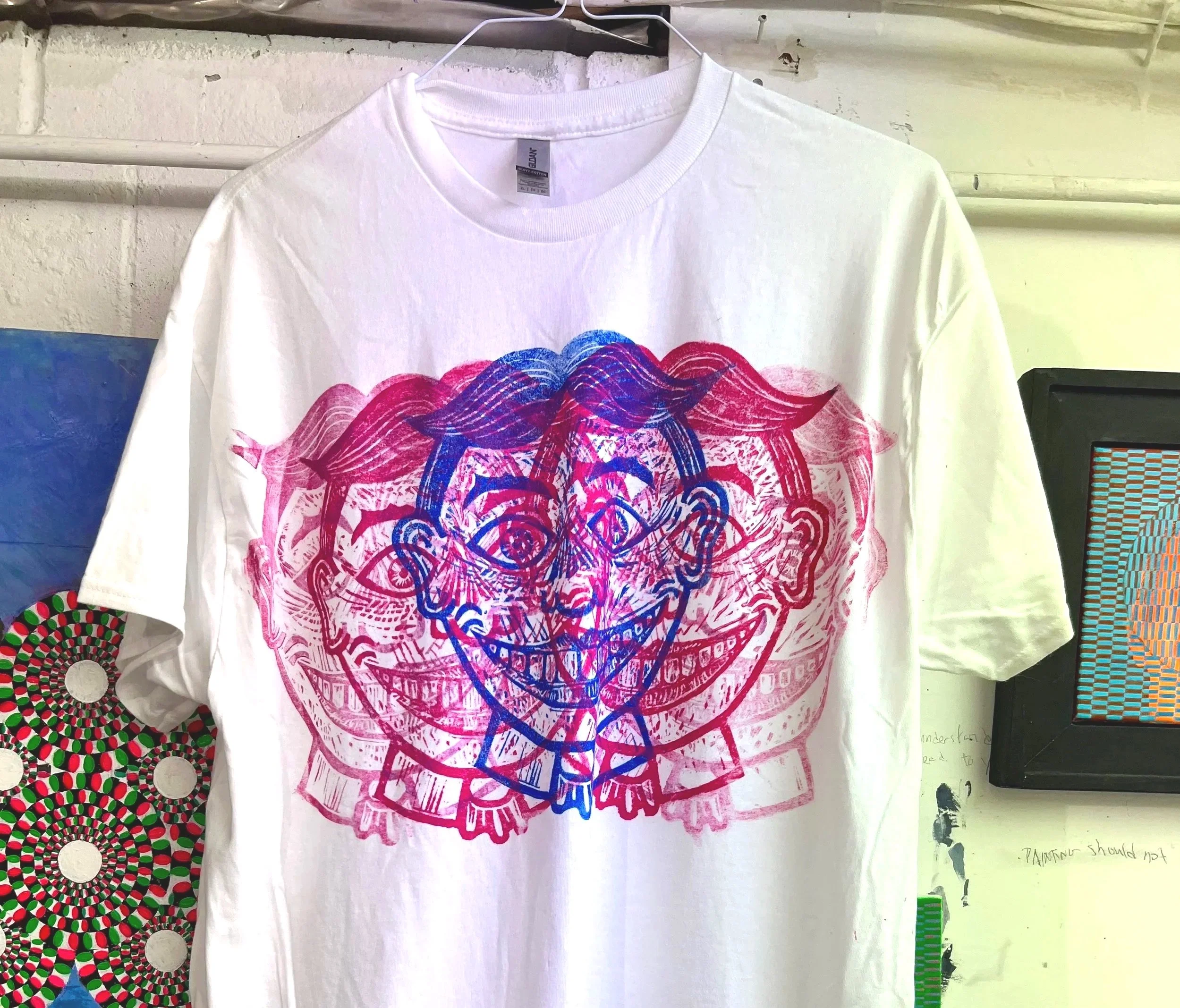 White T-shirt with a colorful, overlapping graphic of a smiling cartoon face with multiple eyes in pink and blue tones hanging on a hanger.