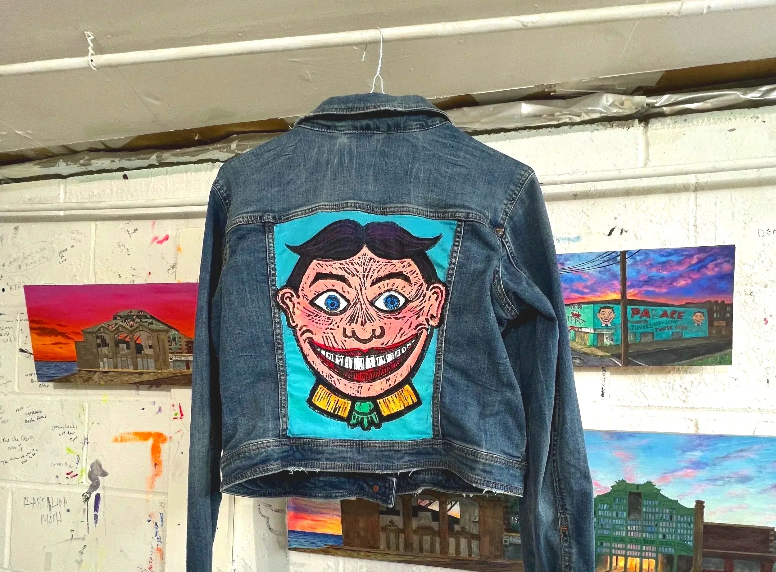 A denim jacket with a colorful, cartoonish patch of a smiling face with big blue eyes, black hair, and a yellow collar, hanging on a hook in an art studio. The background shows various landscape paintings on a white brick wall.