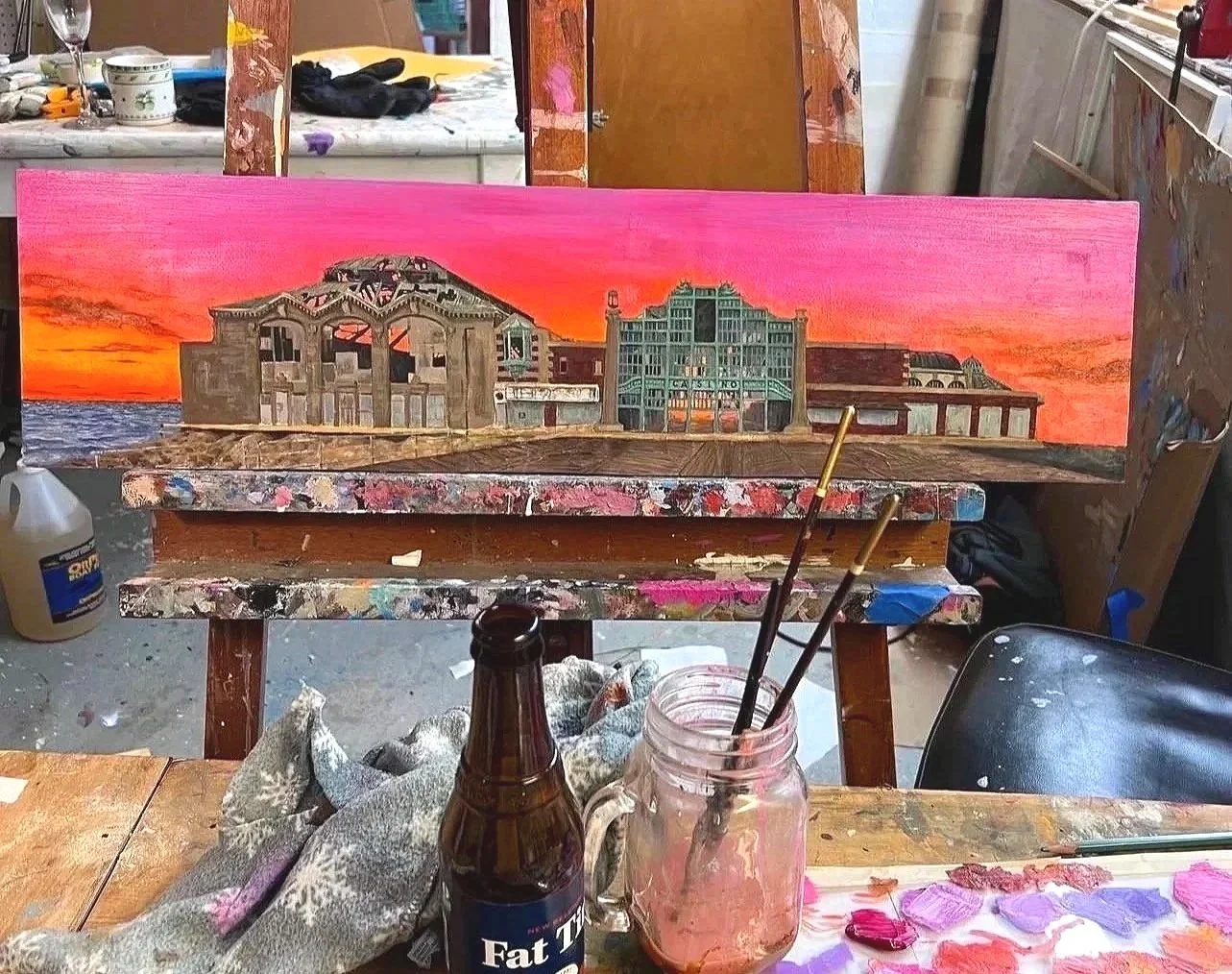A painting in progress on an easel, depicting a coastal scene with a colorful sunset sky, old buildings, and the ocean in the background.