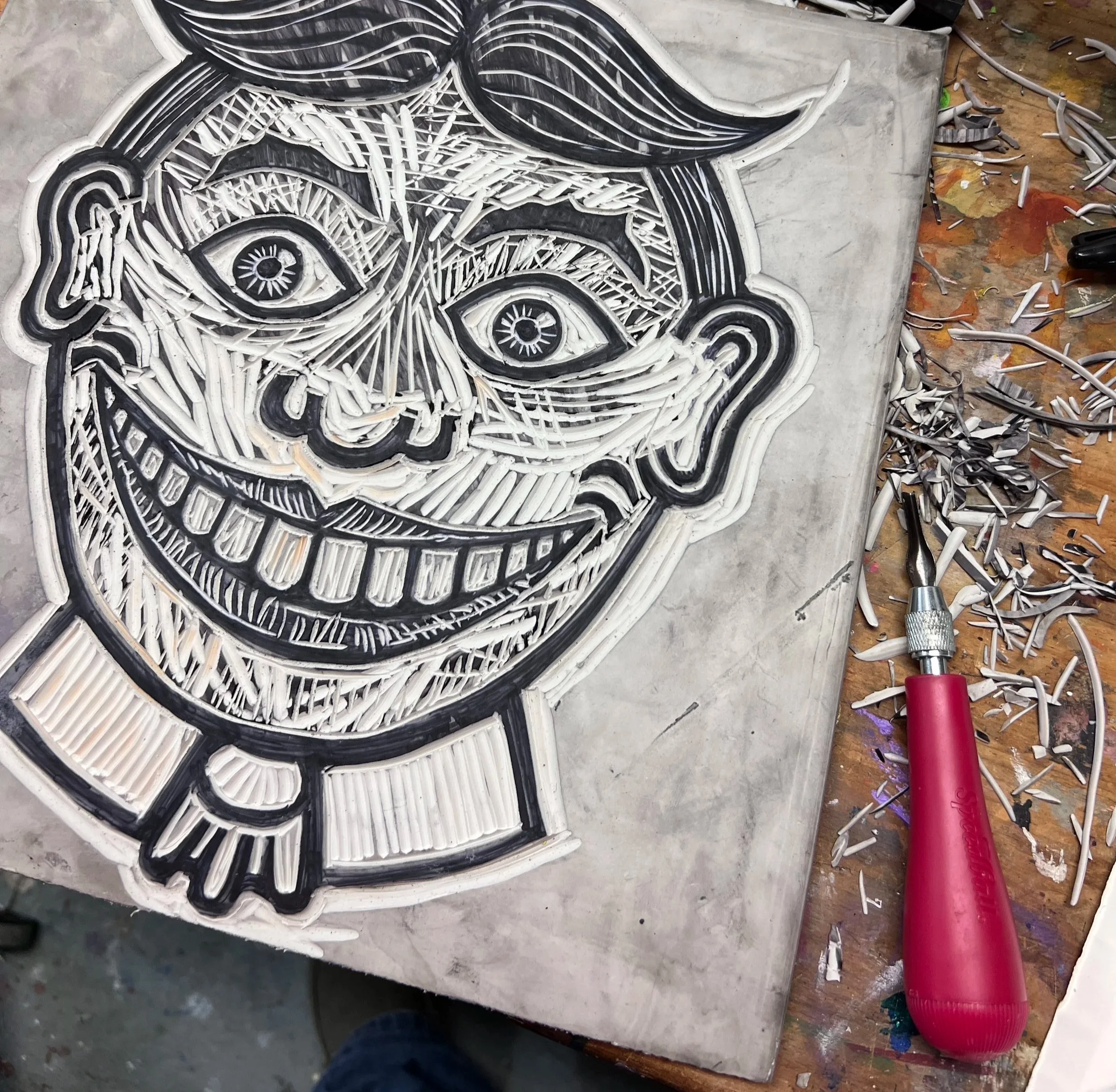 A metal art piece of a smiling face with large eyes, a broad grin, and exaggerated features, created with layered metal strips. A tool with a red handle is on the workbench surrounded by metal shavings.