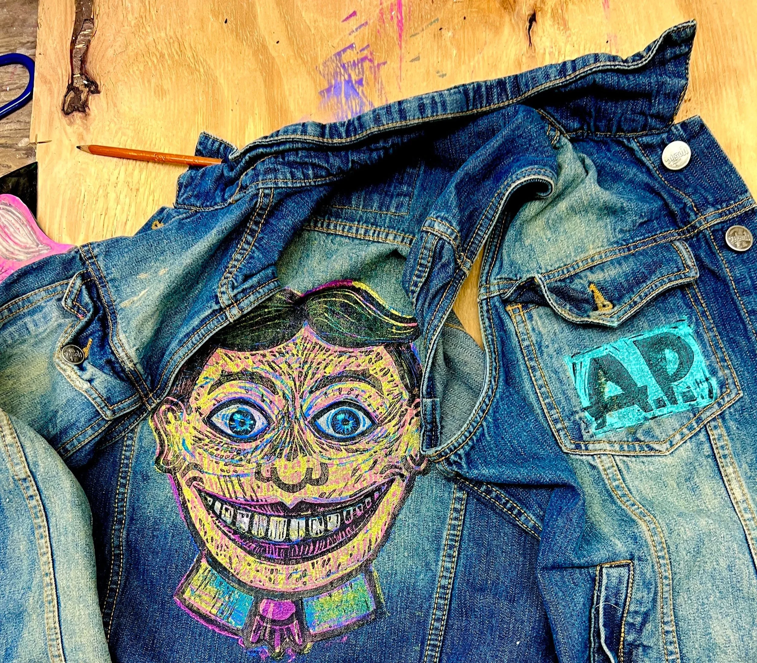 A denim jacket with a colorful, artistic depiction of a person's face on the front, featuring large eyes, a wide smile, and dark hair. The jacket also has a blue patch with the letters 'A.P.' on the back pocket.