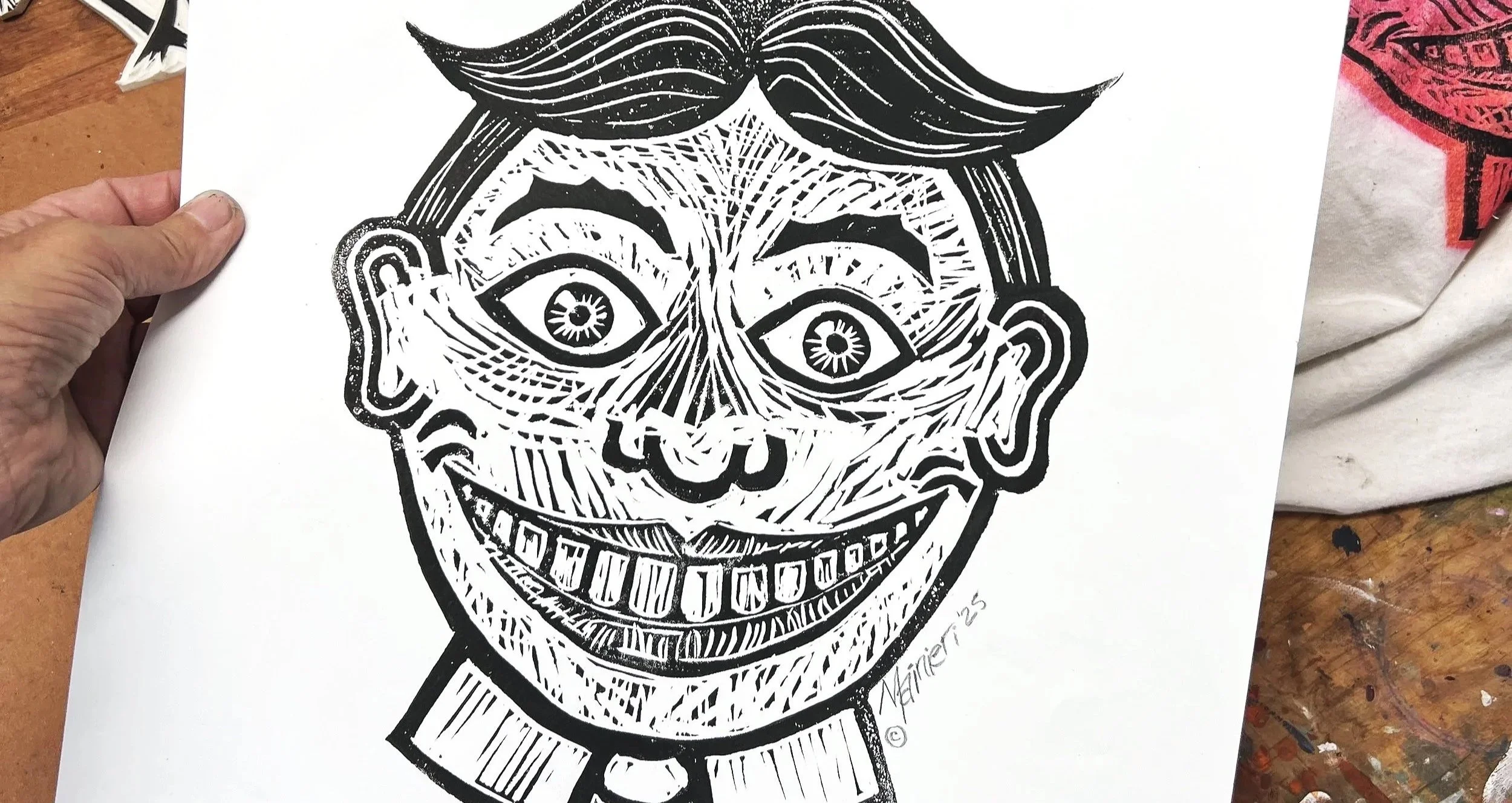 A black and white line art drawing of a person's face with exaggerated features, including three eyes, a wide smile showing teeth, and a mustache, on a white sheet of paper.