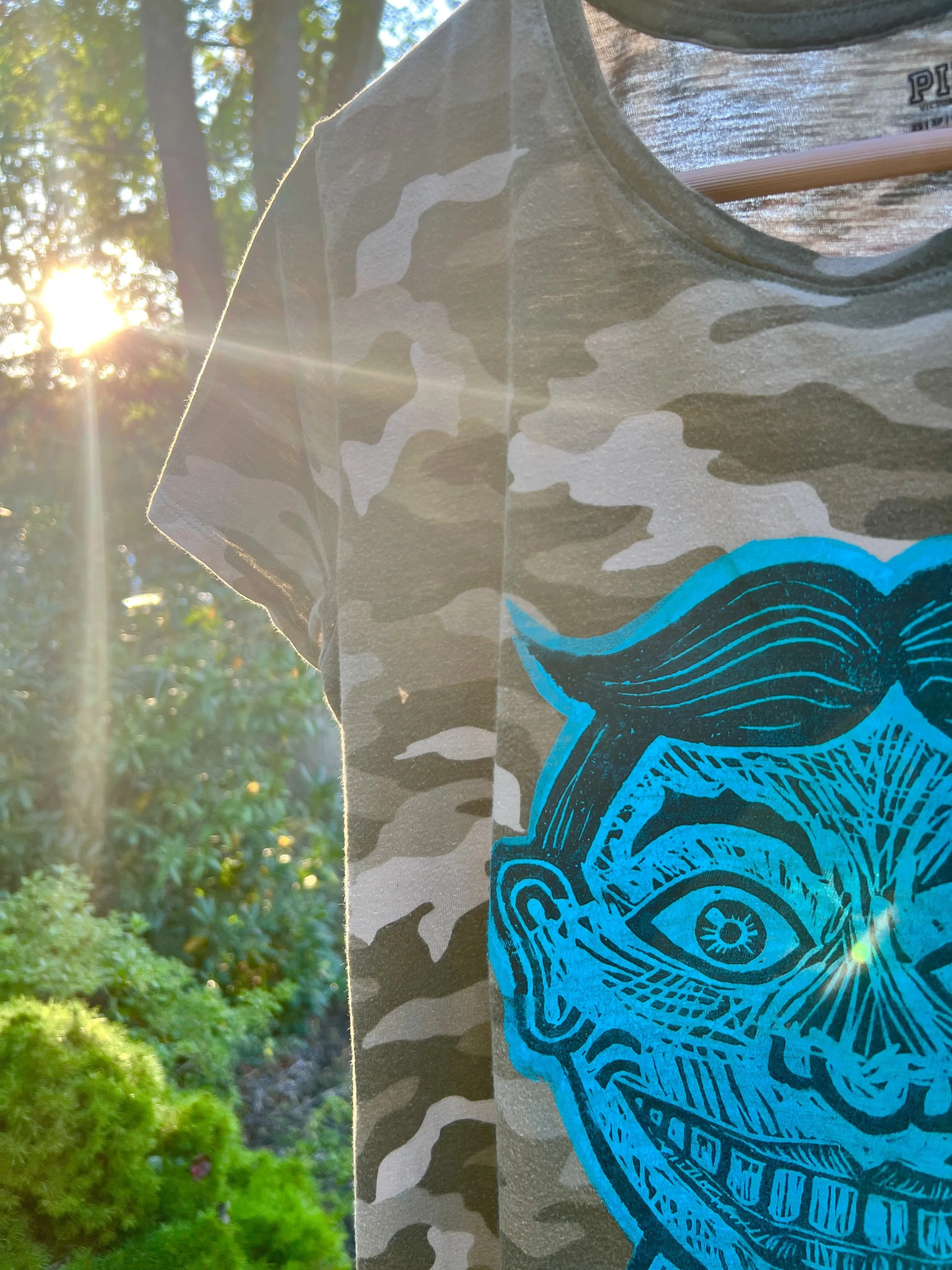 A camouflage T-shirt features a blue graphic of a person's face with an eye and detailed facial features. The T-shirt is outdoors with sunlight filtering through trees in the background.