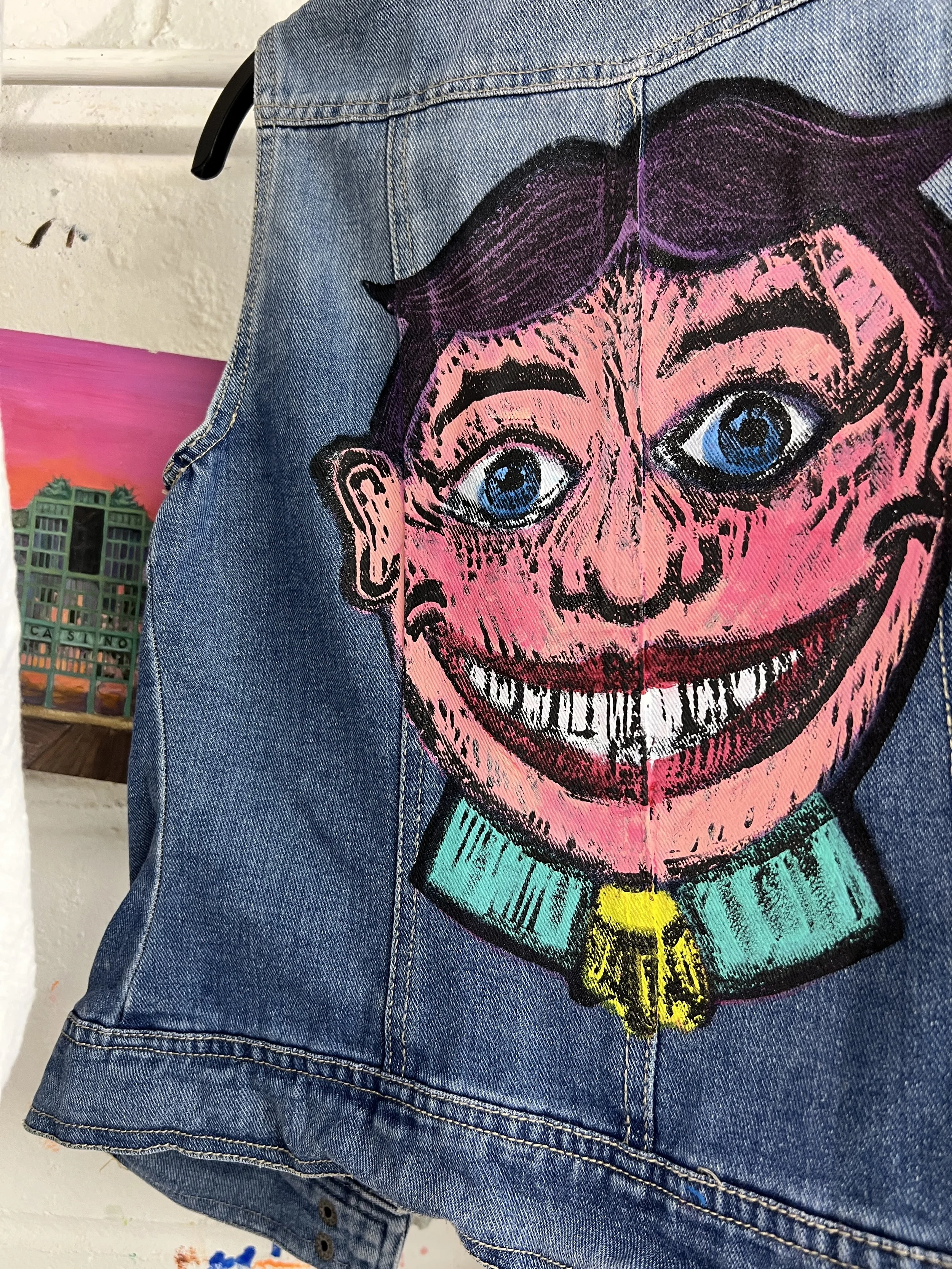 A close-up of a denim jacket featuring a large, colorful painted Tillie from Asbury Park with exaggerated features, including big blue eyes, a wide smiling mouth with white teeth, dark hair, and a green collar with a yellow badge.