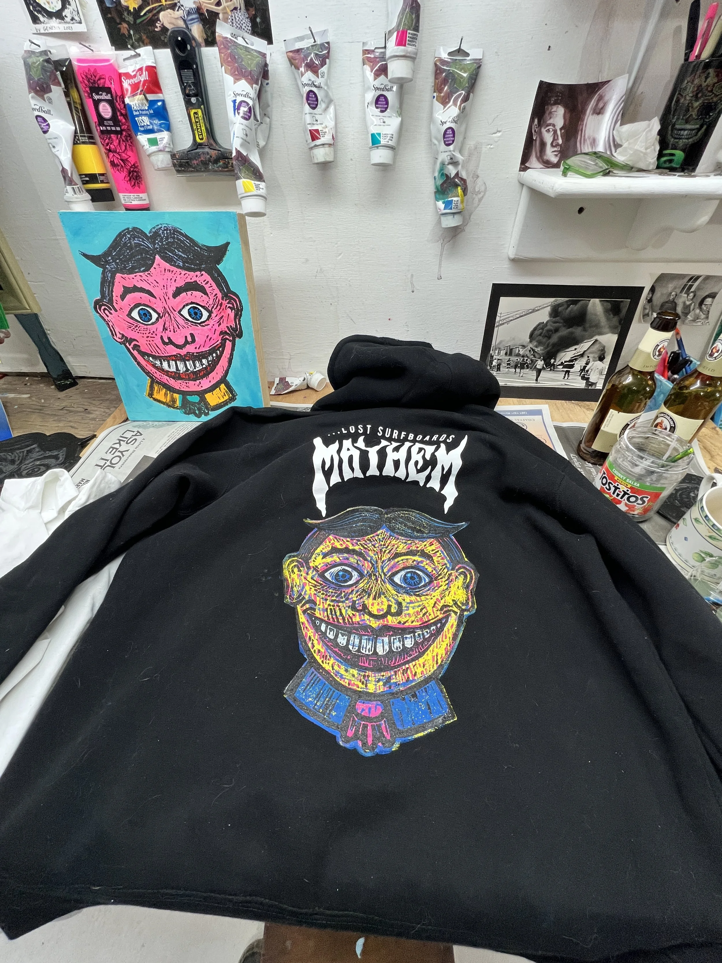 A black hoodie with a colorful, cartoonish portrait of Tillie with blue eyes, dark hair, and a yellow mustache, on a cluttered table surrounded by art supplies, a painting of a pink-faced cartoon character, and some framed photos.