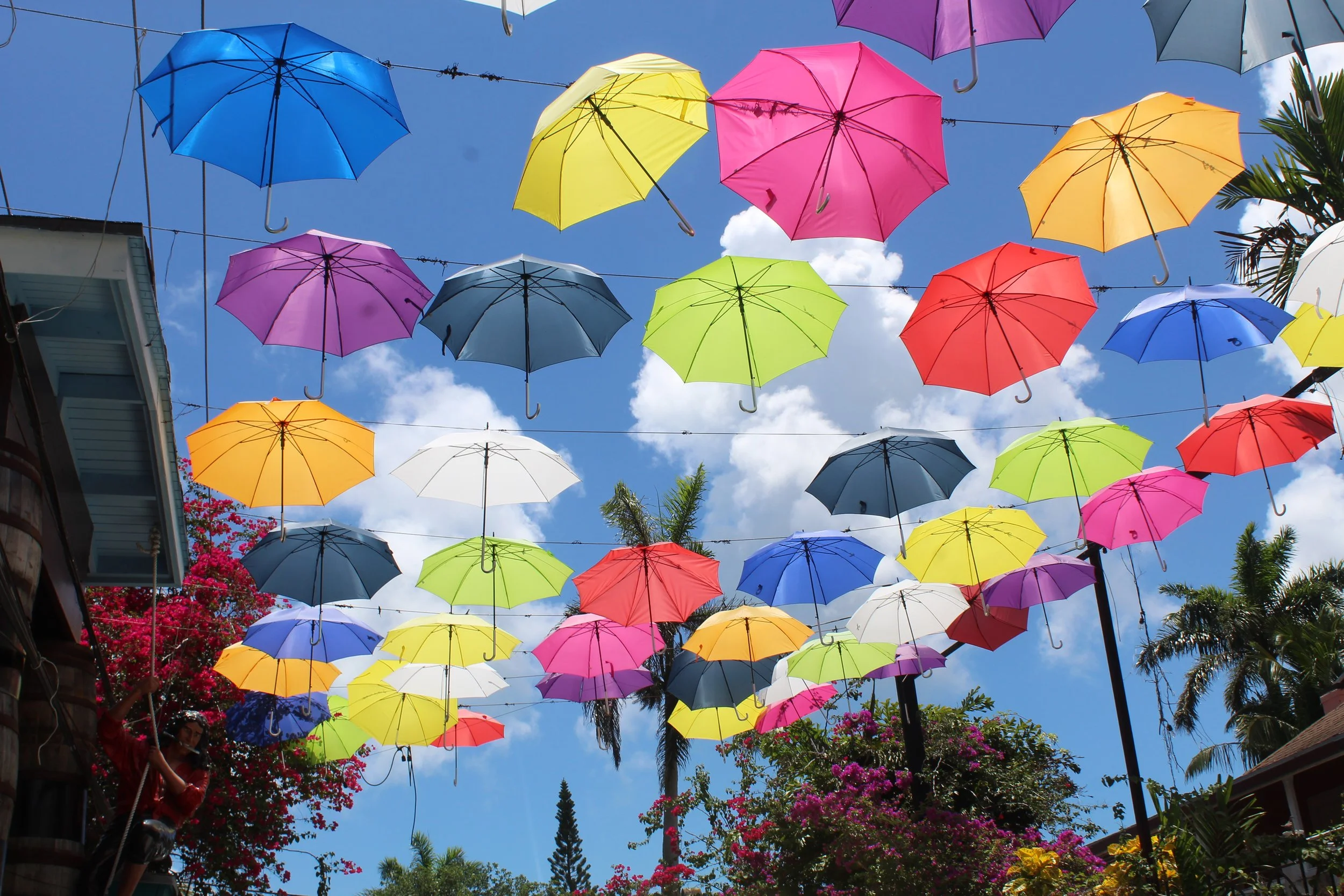 Colorful umbrellas hanging in the sky during daytime, with blue sky and clouds in the background.