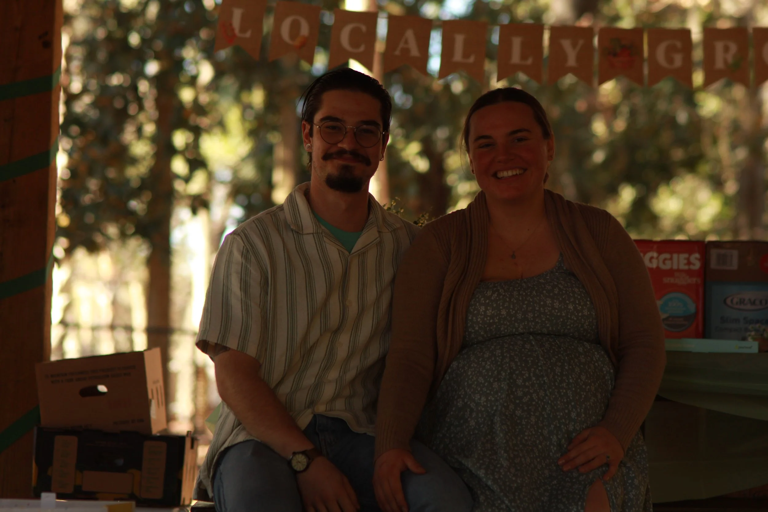 Two smiling people, a man with glasses and a woman, posing together at an outdoor event with trees in the background and a banner that says 'Locally Grown' hanging above their heads.