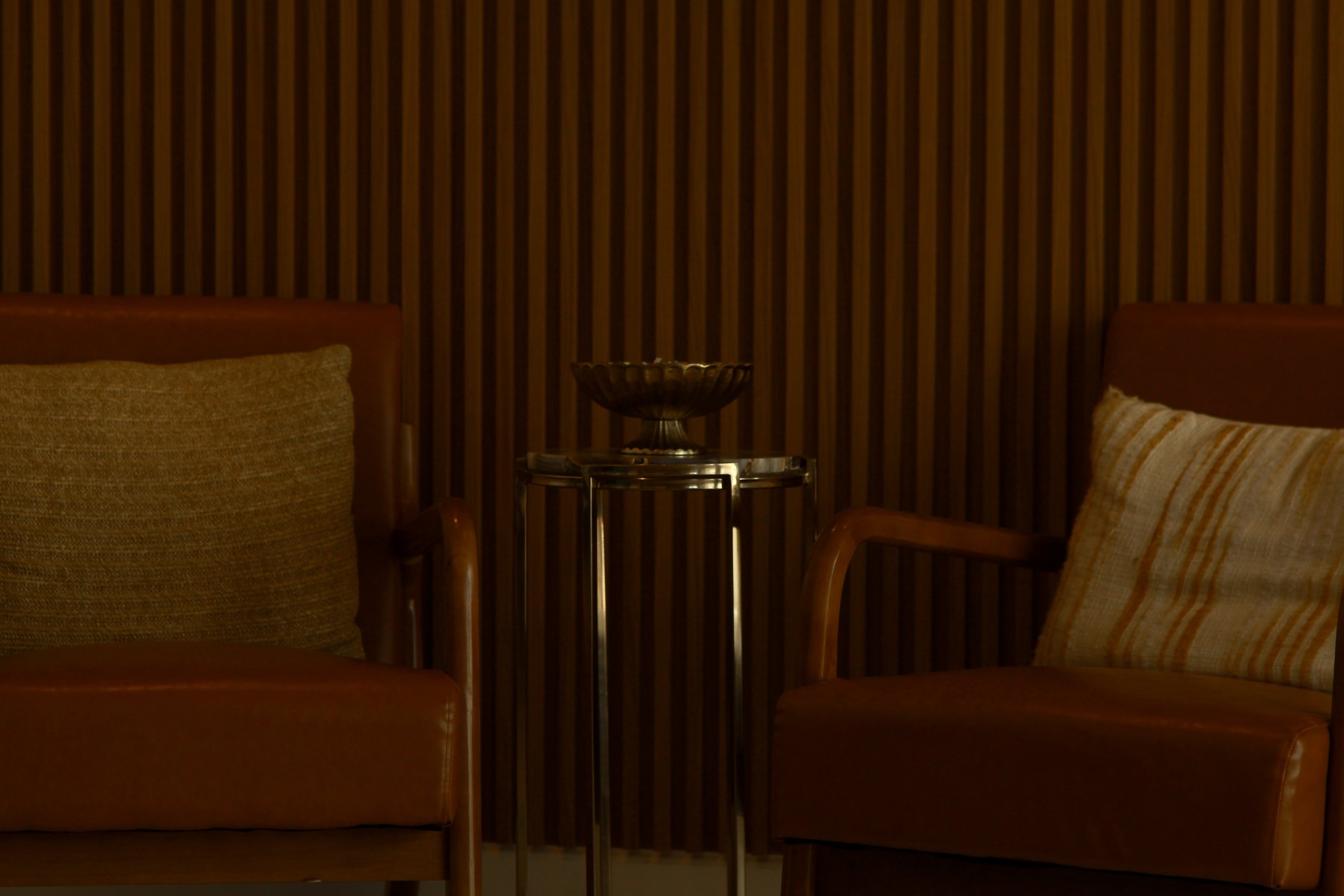 Two leather armchairs with cushions and a small table between them in a dimly lit room with wooden paneling on the wall.