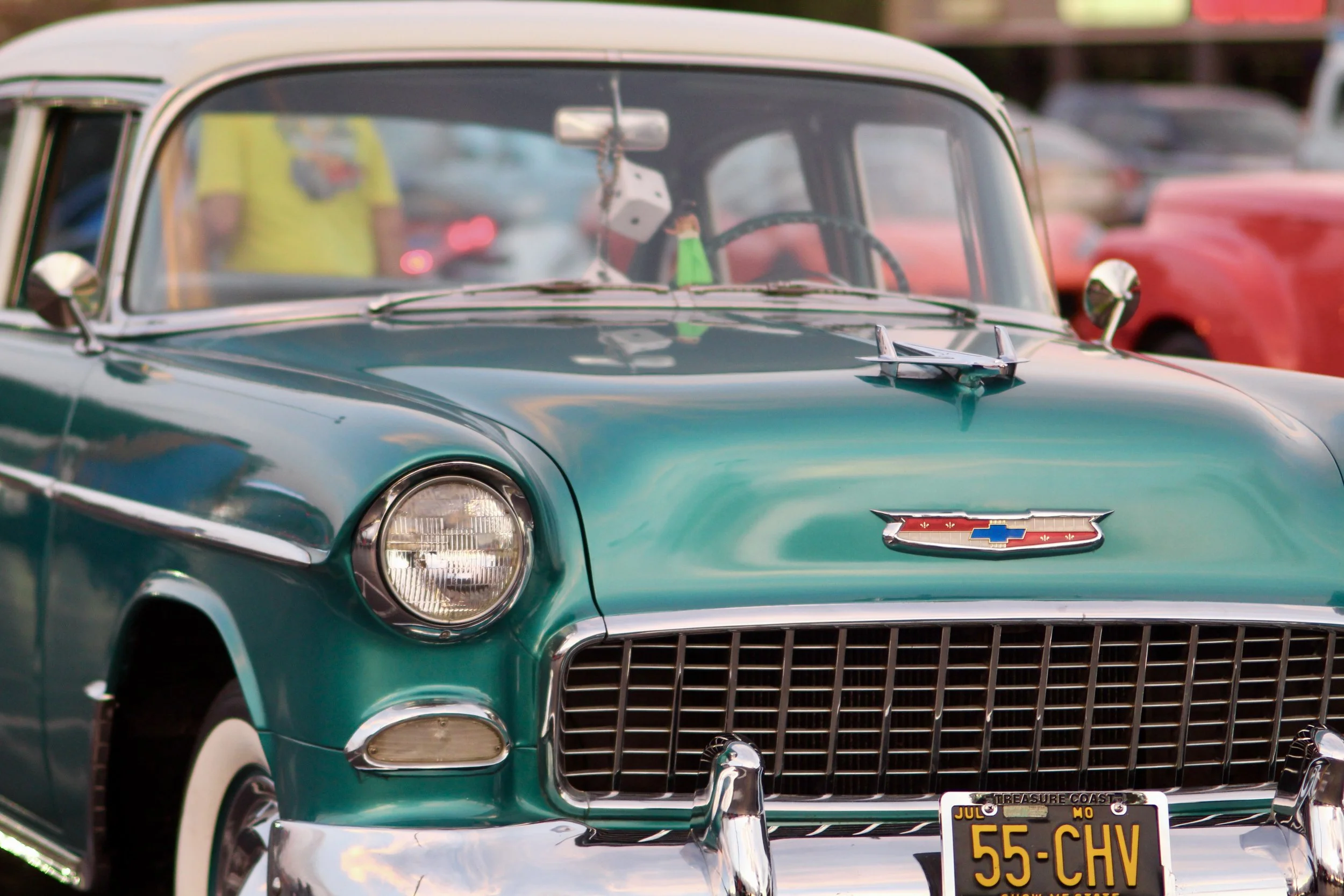 A vintage teal Chevrolet car on display, featuring a front grille and a license plate with the text '55-CHV'. The car has a chrome bumper and a Chevrolet emblem on the front.