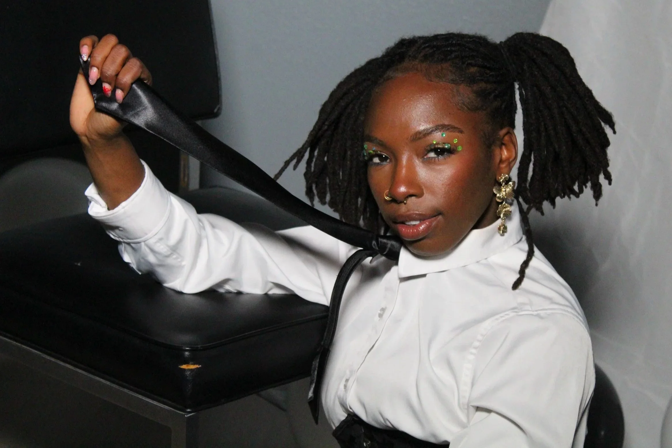 A woman with dreadlocks adorned with green beads and earrings, sitting in front of a black speaker, holding a black strap with her right hand, wearing a white shirt.