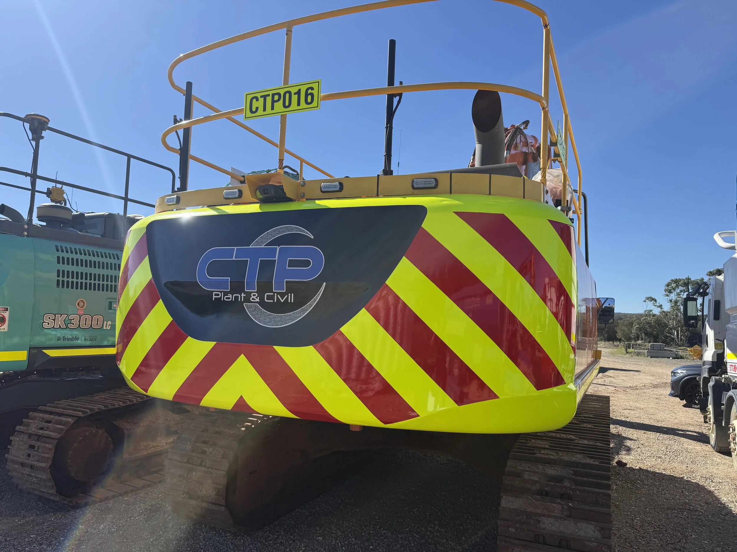 A construction excavator with yellow and red striped safety markings, labeled CPT16, and a logo that reads 'CTP Plant & Civil.' It has a black arm and tracks, and is parked outdoors with other construction equipment and vehicles visible in the background under a clear blue sky.