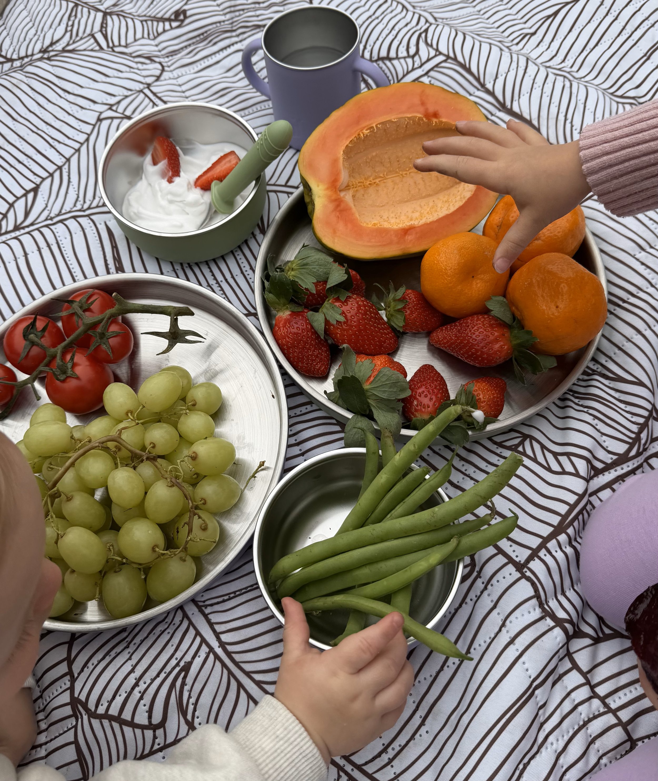 A variety of fruits including green grapes, cherry tomatoes, strawberries, a halved cantaloupe, oranges, and a bunch of green beans are displayed on a patterned tablecloth. Two hands are reaching toward the fruits.
