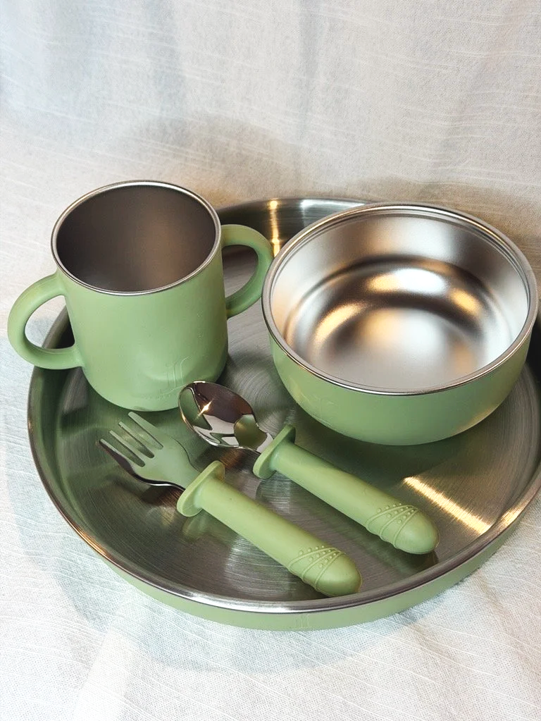 A green metal tray holding a green mug, a green bowl, a green fork, and a green spoon, all with metal interiors, on a light-colored surface.