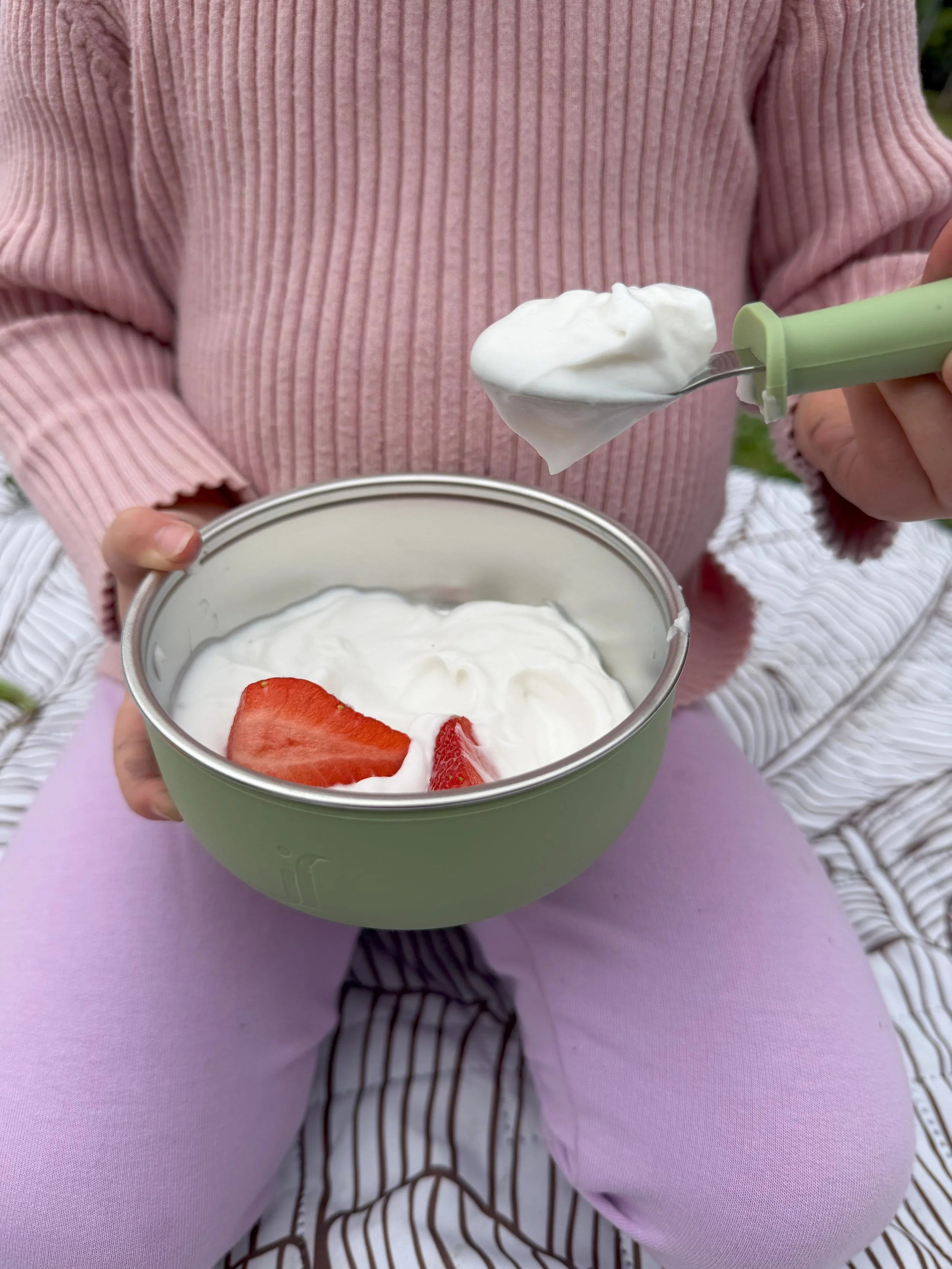 Person holding a spoon with yogurt and strawberry slices in a light green bowl, sitting outdoors on purple fabric.