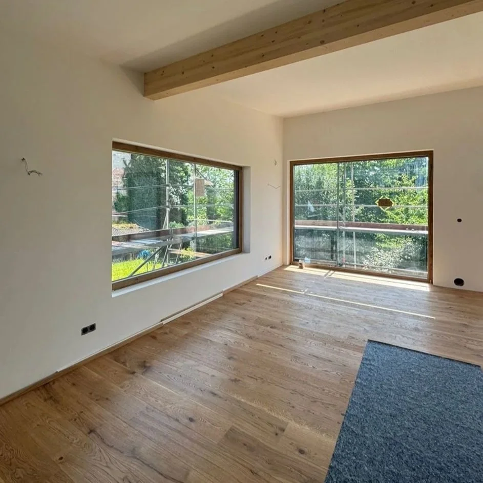 Ein heller, leerer Raum mit großen Fenstern, frisch verlegt Holzboden und einer sichtbaren Holzbalken-Decke.