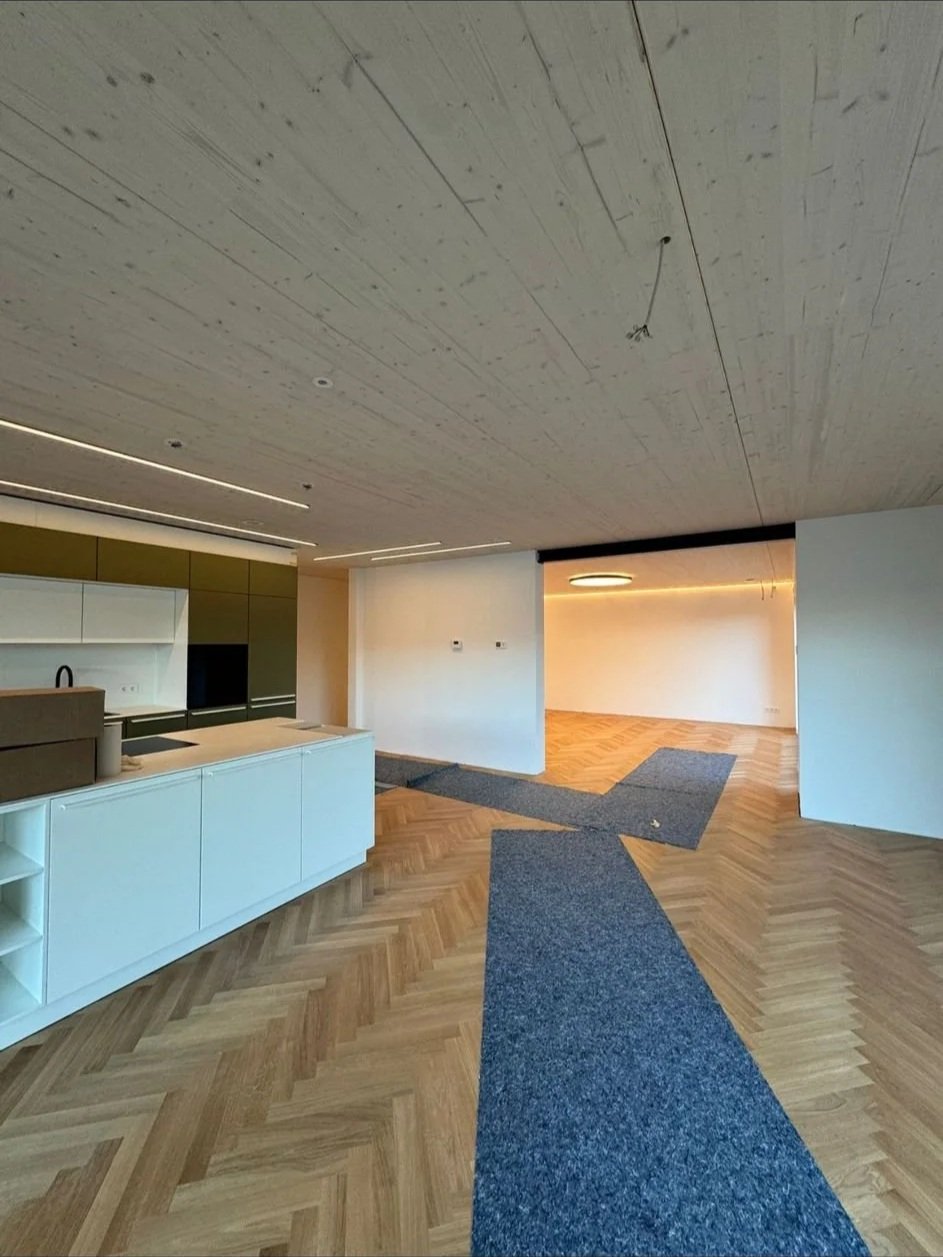 Innenraum eines modernen, unfertigen Apartments mit Holzparkett, weißen Wänden und einer offenen Küche mit weißen und grünen Schränken. Der Boden ist teilweise mit blauen Teppichen bedeckt.