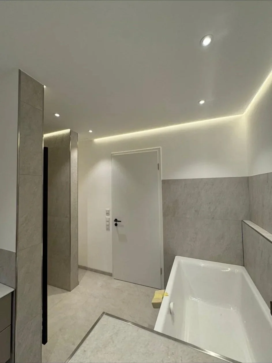 Moderne Badezimmer mit beigen Fliesen, einer weißen Tür, einer Badewanne und integrierter Deckenbeleuchtung.