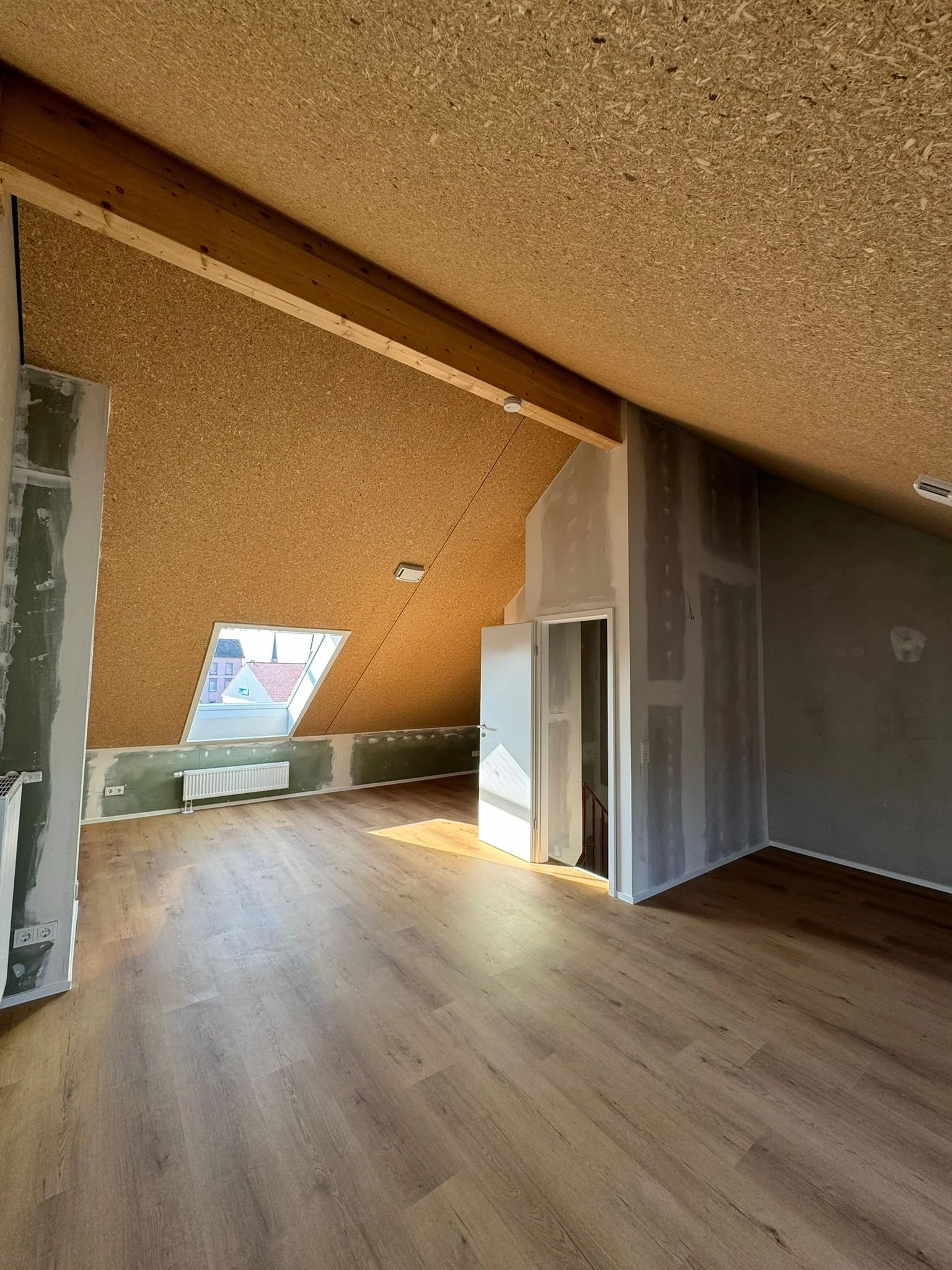 Ein unmöbliertes Zimmer mit Holzboden, schrägem Dach und einem großen Fenster auf der Dachseite, das Tageslicht hereinlässt.