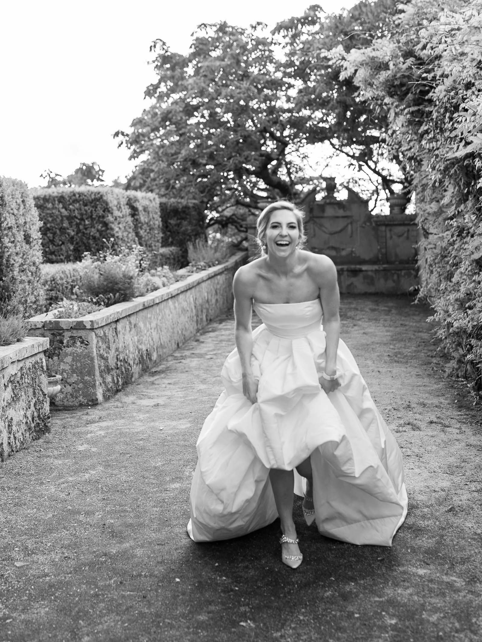 A joyful bride in a strapless wedding gown, lifting the front of her skirt to reveal her shoes, standing on a garden pathway surrounded by trees and bushes.