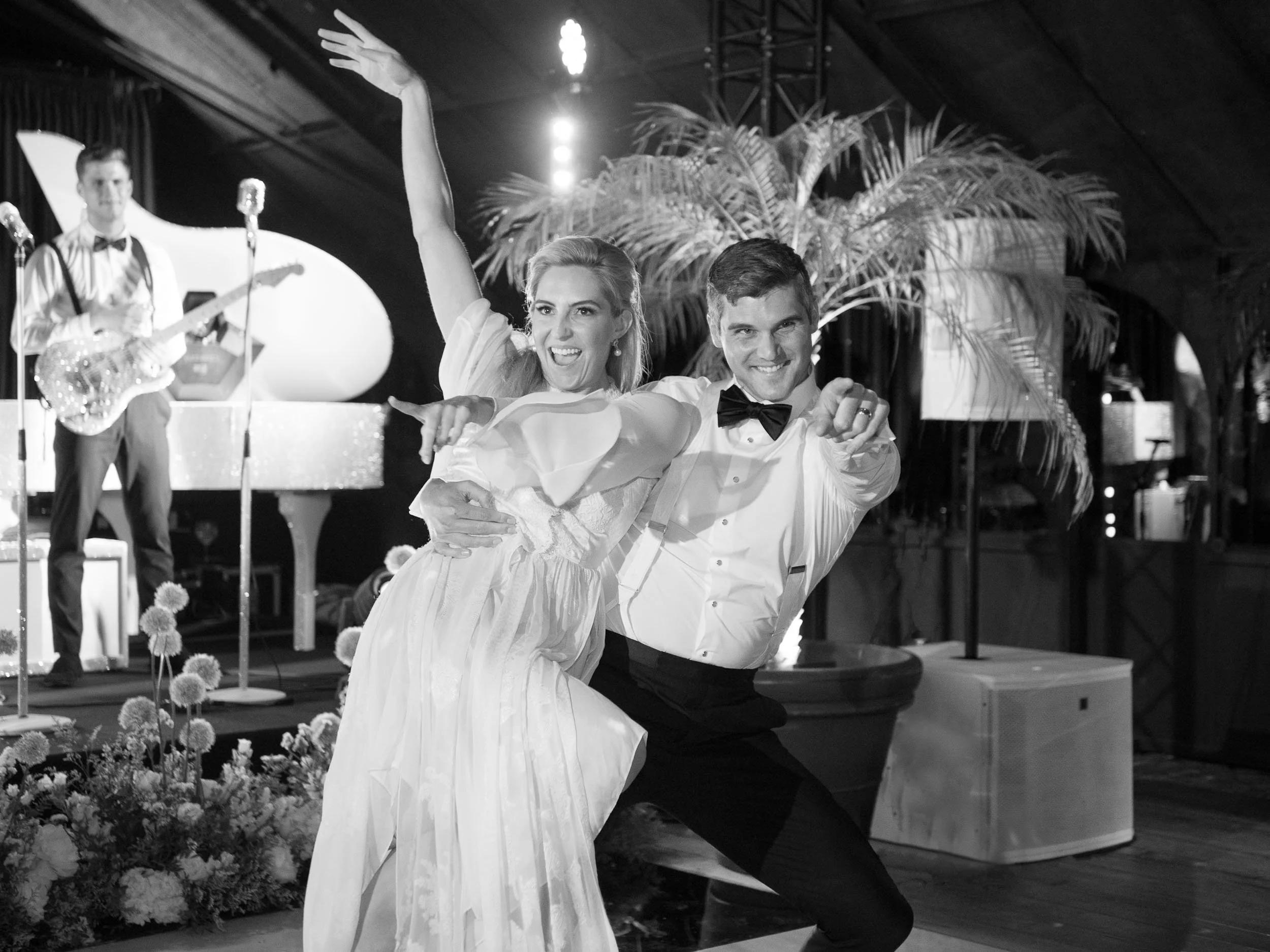A wedding reception with a bride and groom dancing and posing playfully. The bride is wearing a white wedding dress, and the groom is in a tuxedo with a bow tie. In the background, a jazz band is playing, including a guitarist and a singer, with deco