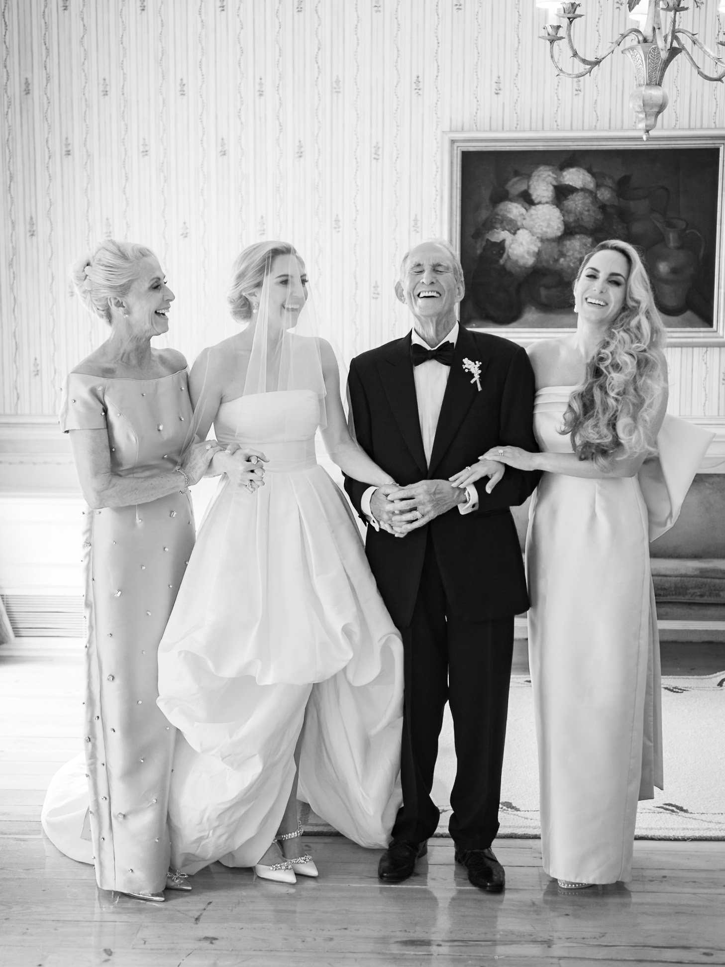 Black and white photo of  women and one man, all smiling and holding hands, standing in an elegant room. The woman in the center is dressed as a bride, wearing a wedding gown and veil. The man, likely the father of the bride, is wearing a tuxedo. The