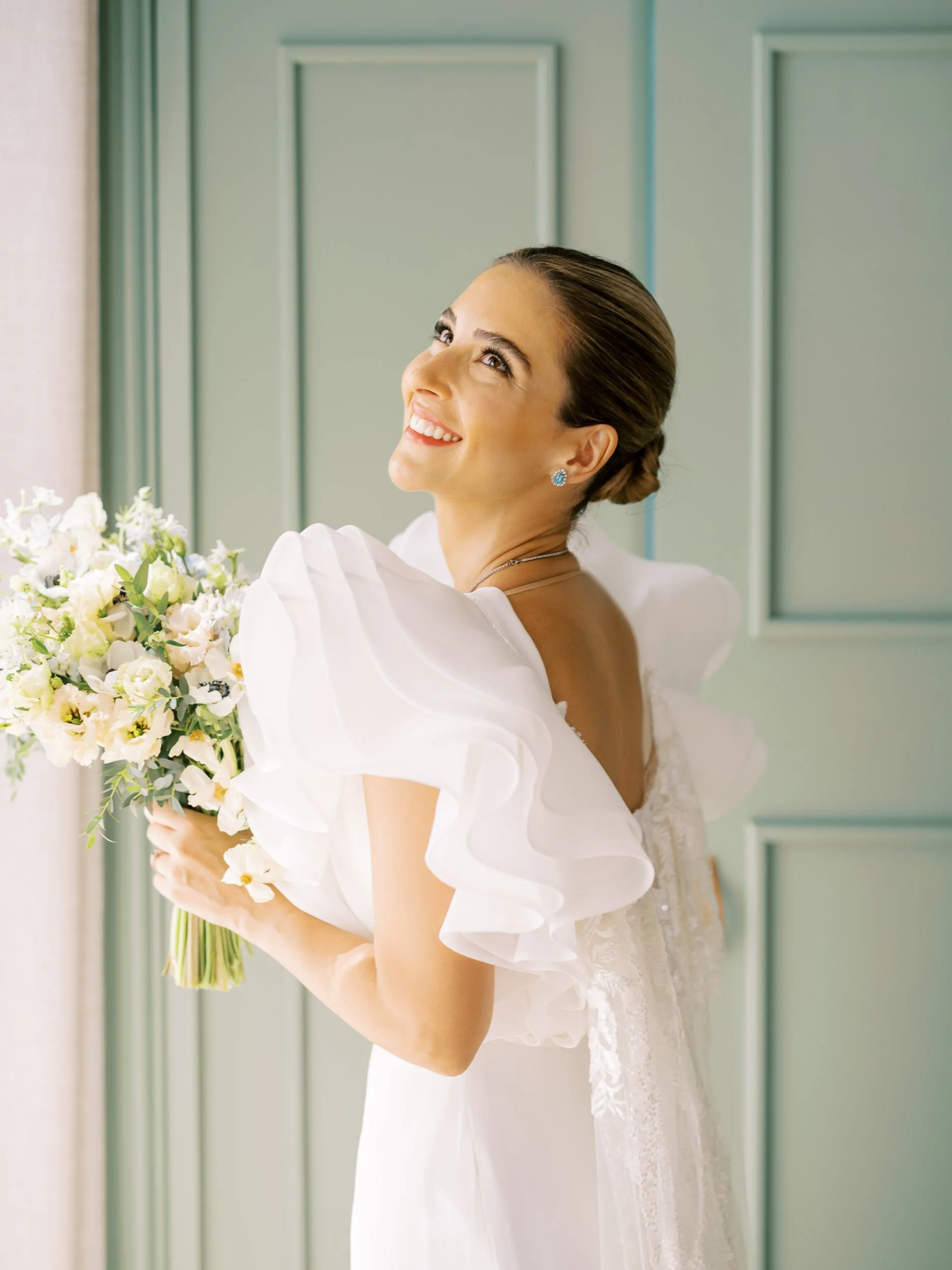A smiling bride in a white dress holding a bouquet of white flowers, standing in front of green paneled doors.