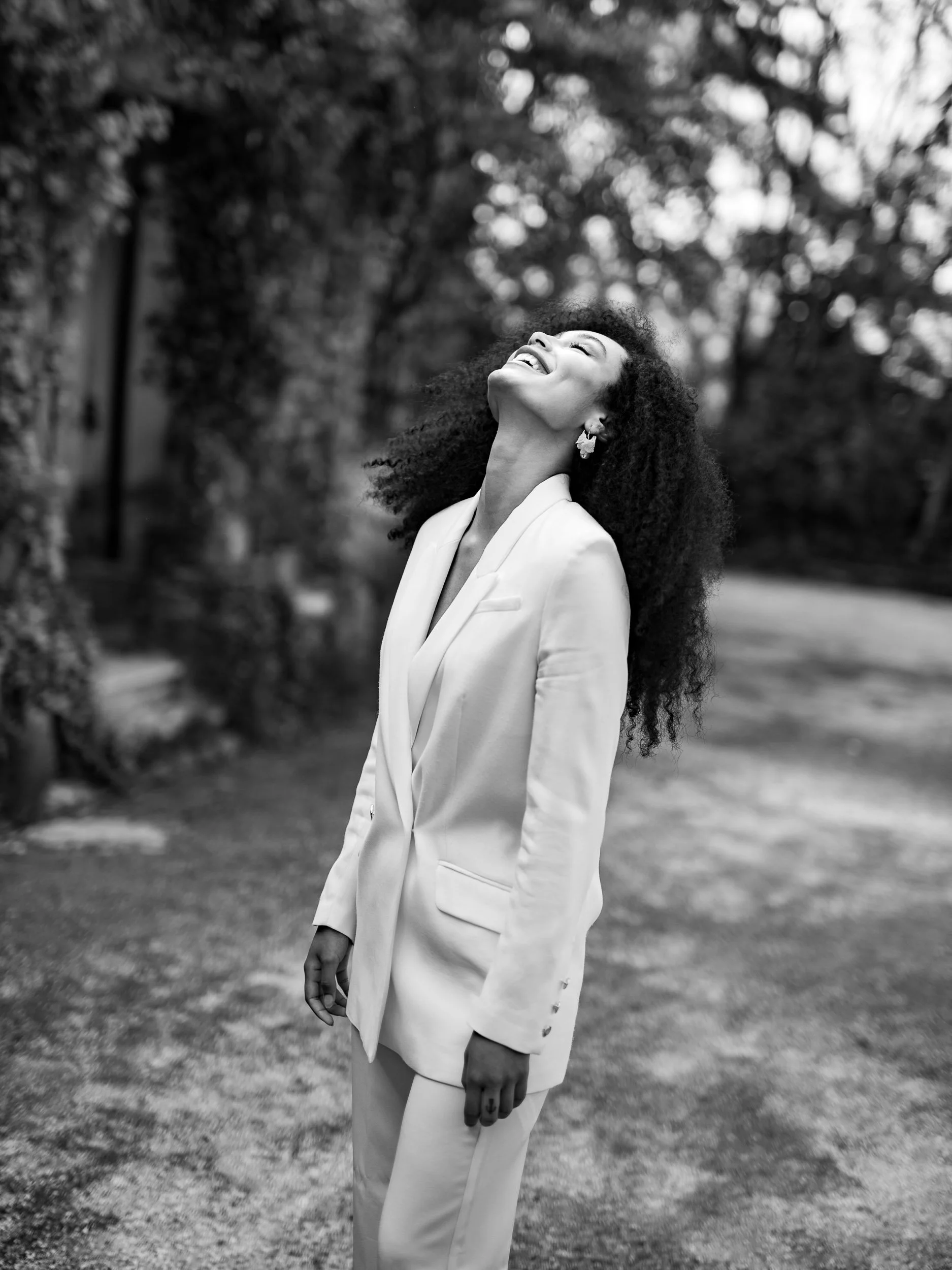 A bride with curly hair smiling and looking up while wearing a white suit outdoors, in black and white.