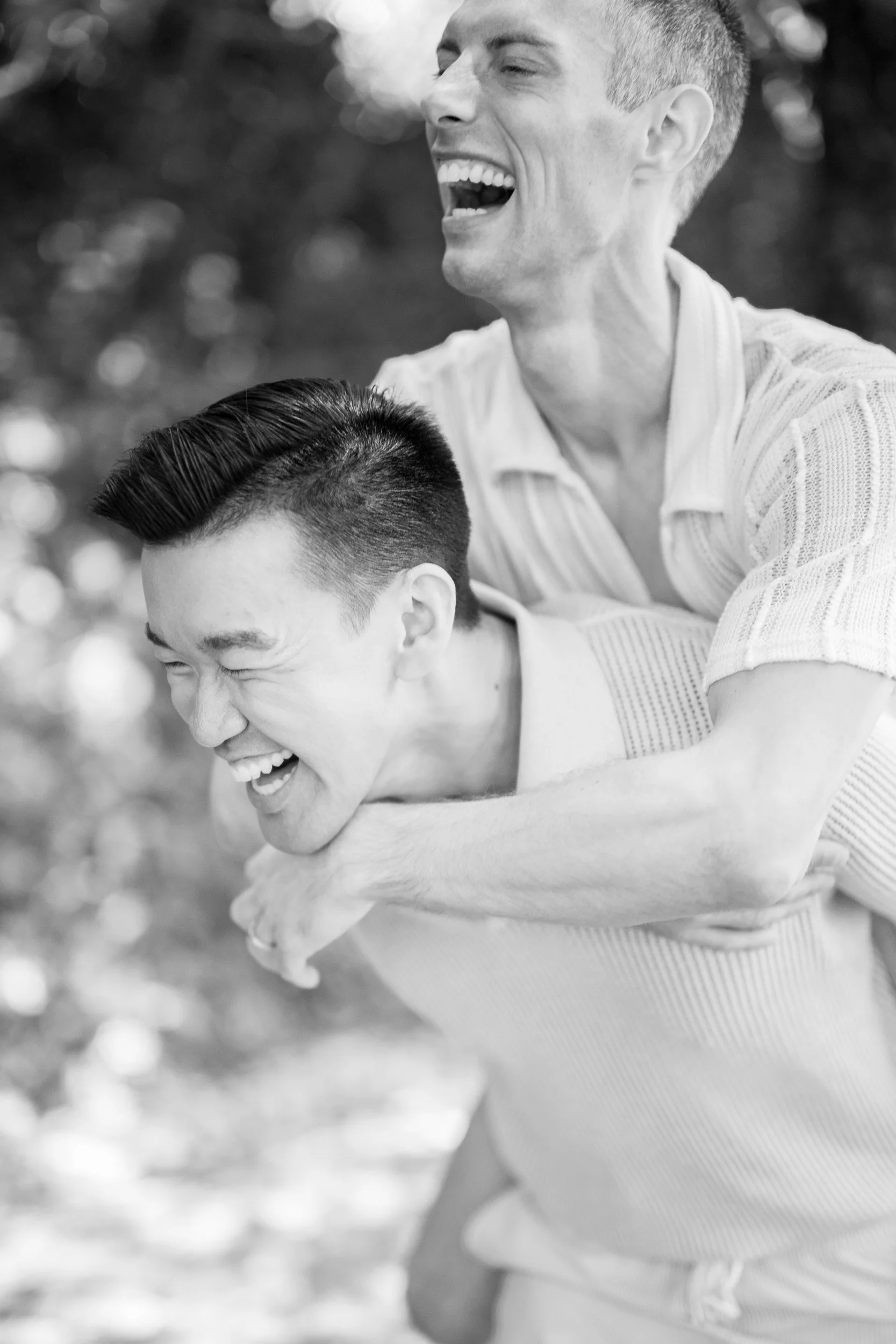 Two grooms laughing and having fun outdoors, one giving the other a piggyback ride.