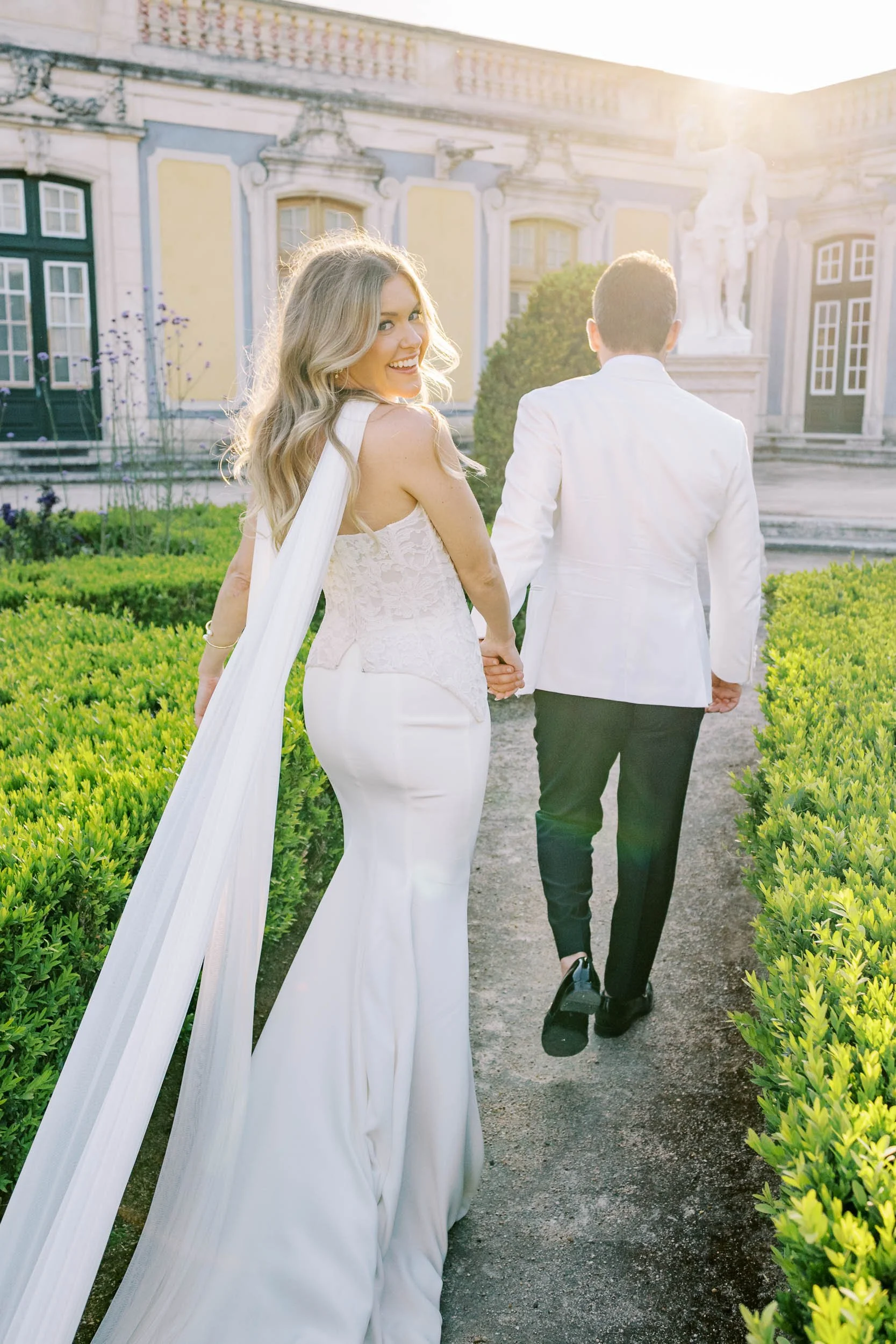 A bride and groom holding hands and walking away on a garden path, with the bride smiling and looking back at the camera, in front of a historic building and statues, illuminated by warm sunlight at Palácio de Queluz in Lisbon