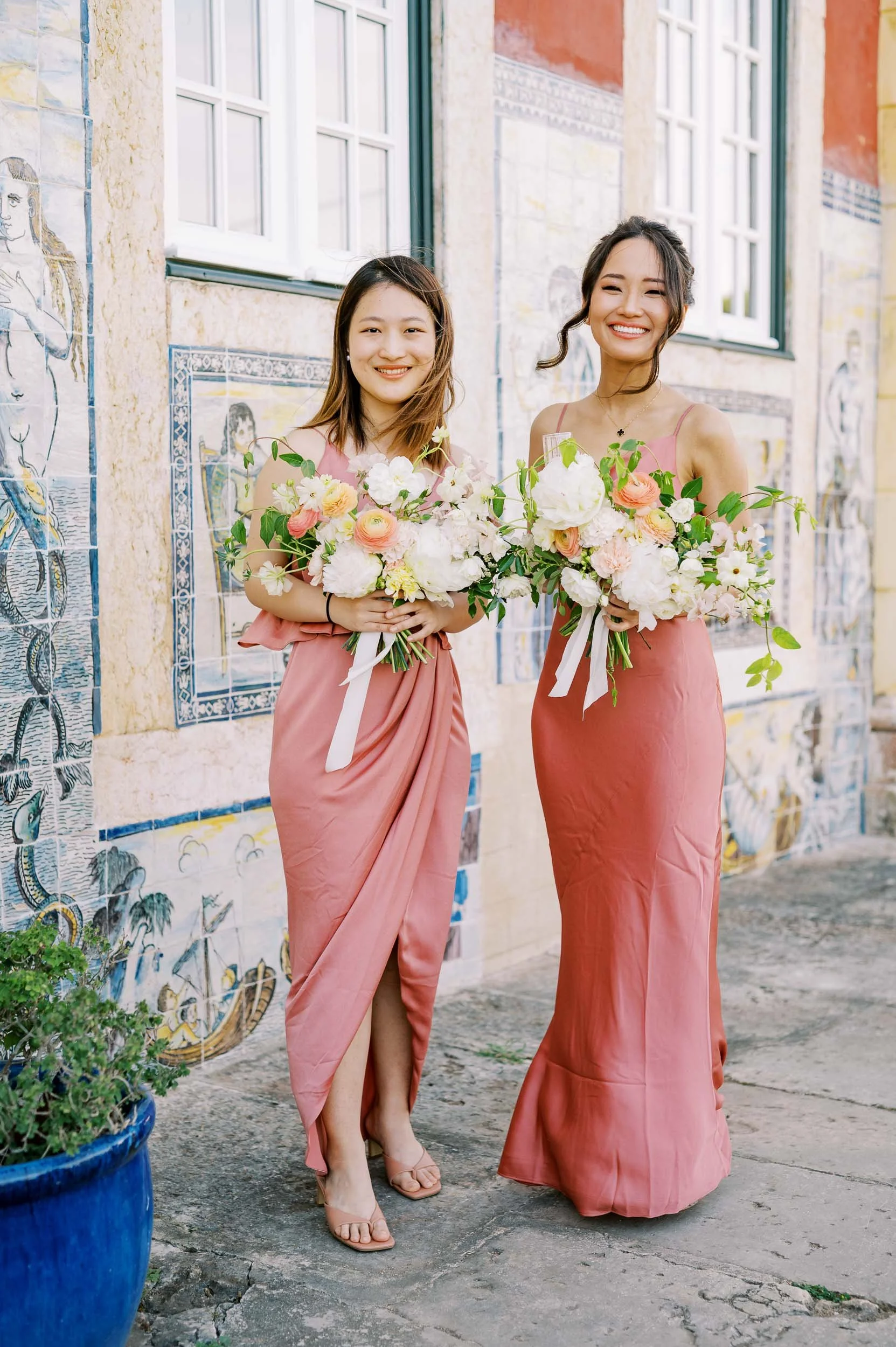 Two bridesmaids in pink dresses holding large bouquets of flowers, standing in front of a colorful mural wall with windows.