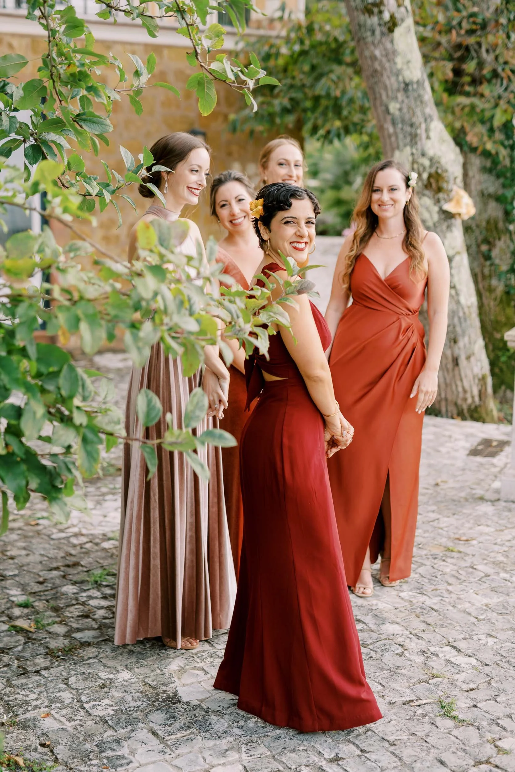 A group of bridesmaids dressed in elegant dresses, standing outdoors on a cobblestone path with trees and greenery in the background, smiling and posing for a photo at Quinta da Bella Vista in Sintra