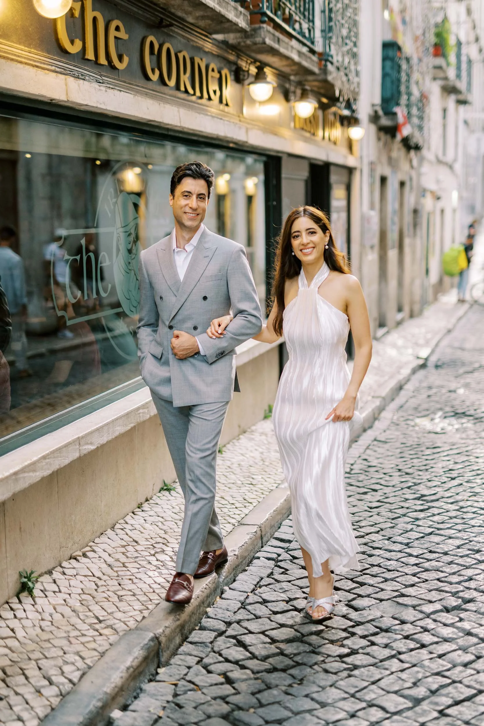 A smiling couple dressed in formal attire walking arm in arm down a cobblestone street outside a restaurant named 'The Corner' during an engagement photo session in Lisbon.
